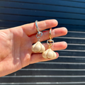 A hand holding a pair of garlic-shaped drop keychains, one with a silver-tone top and the other with a gold-tone top, against a dark blue slatted background.
