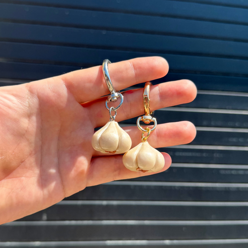A hand holding a pair of garlic-shaped drop keychains, one with a silver-tone top and the other with a gold-tone top, against a dark blue slatted background.