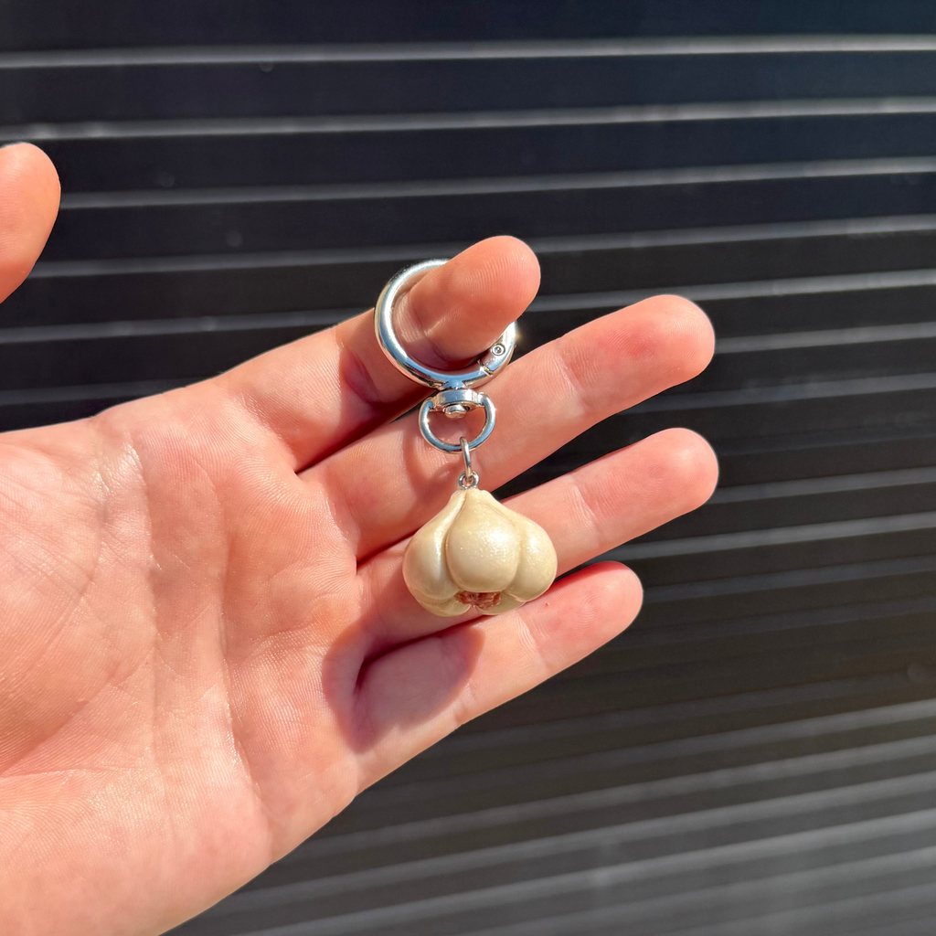 A hand holding a garlic-shaped charm keychain with a silver metal ring and connector against a dark slatted background.