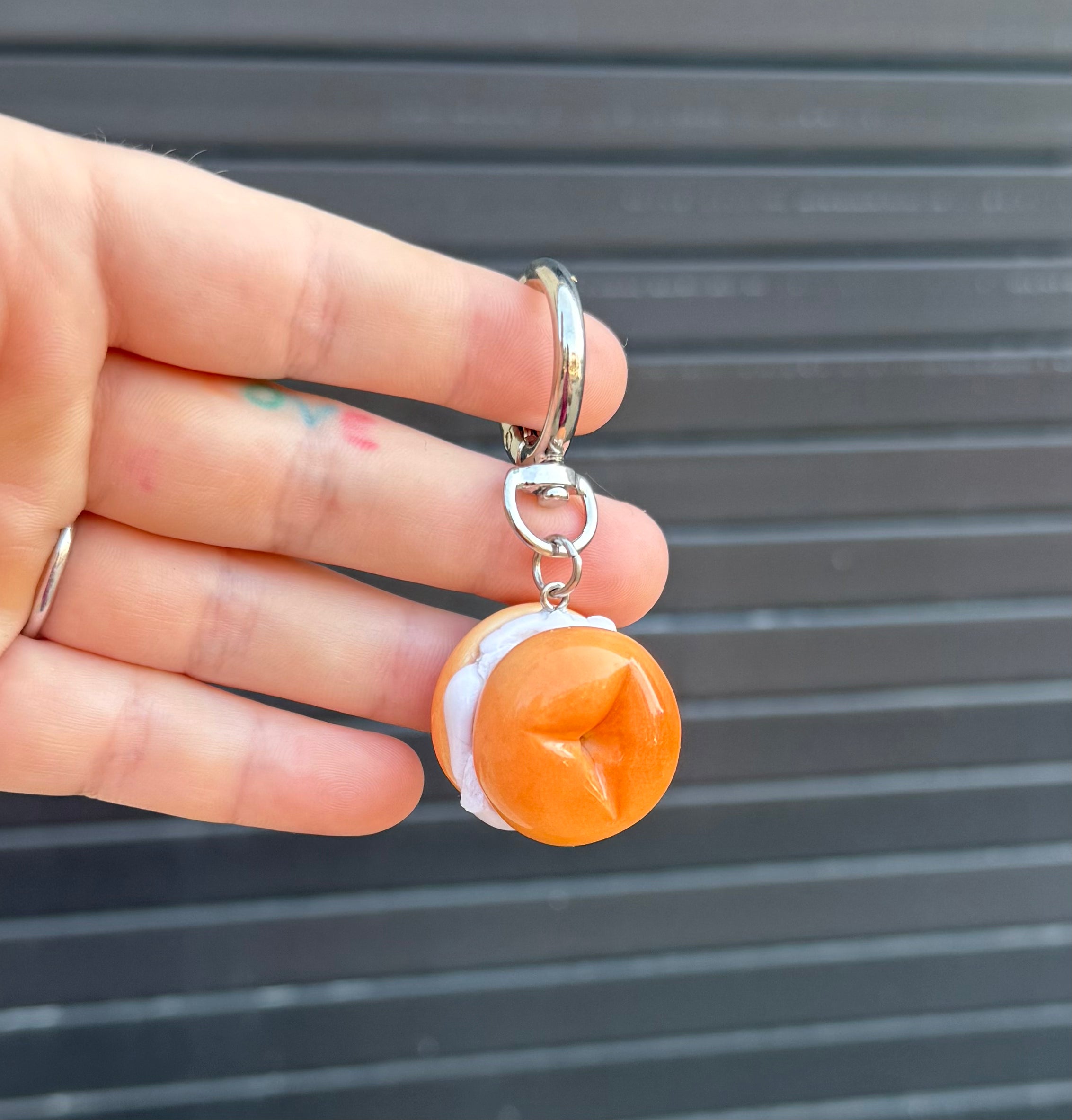 A hand holding a small food-themed keychain charm shaped like a bagel, set against a dark gray slatted background.