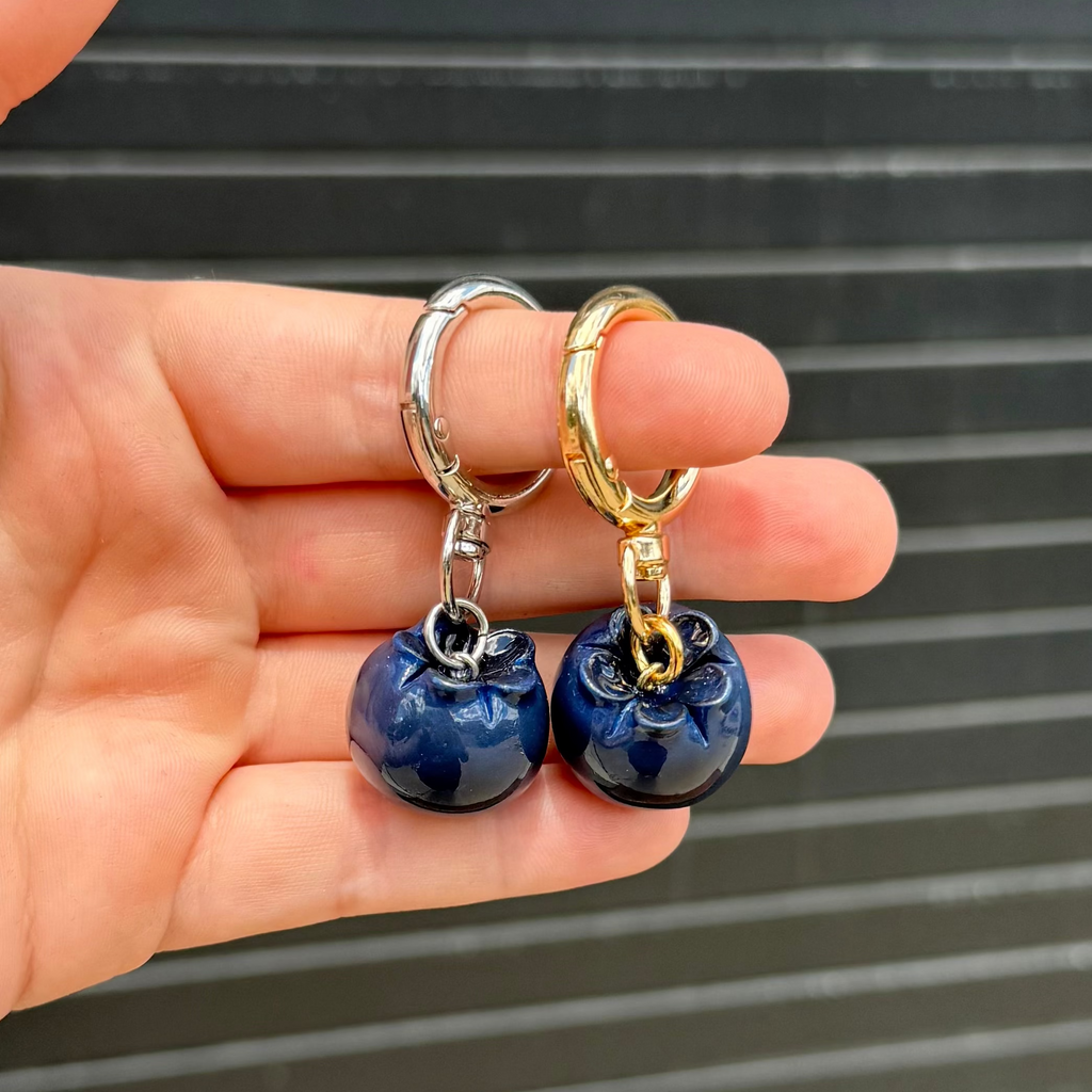 A hand holding two small blueberry keychains, one with a silver-tone hanger and one with a gold-tone hanger, both with glossy dark blue polymer clay bodies, against a dark gray slatted background.