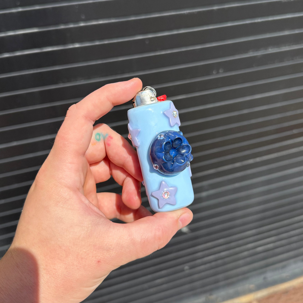 A hand holding a light blue decorative lighter case with a blueberry in the center, purple star embellishments, and a silver top, set against a dark corrugated metal background.