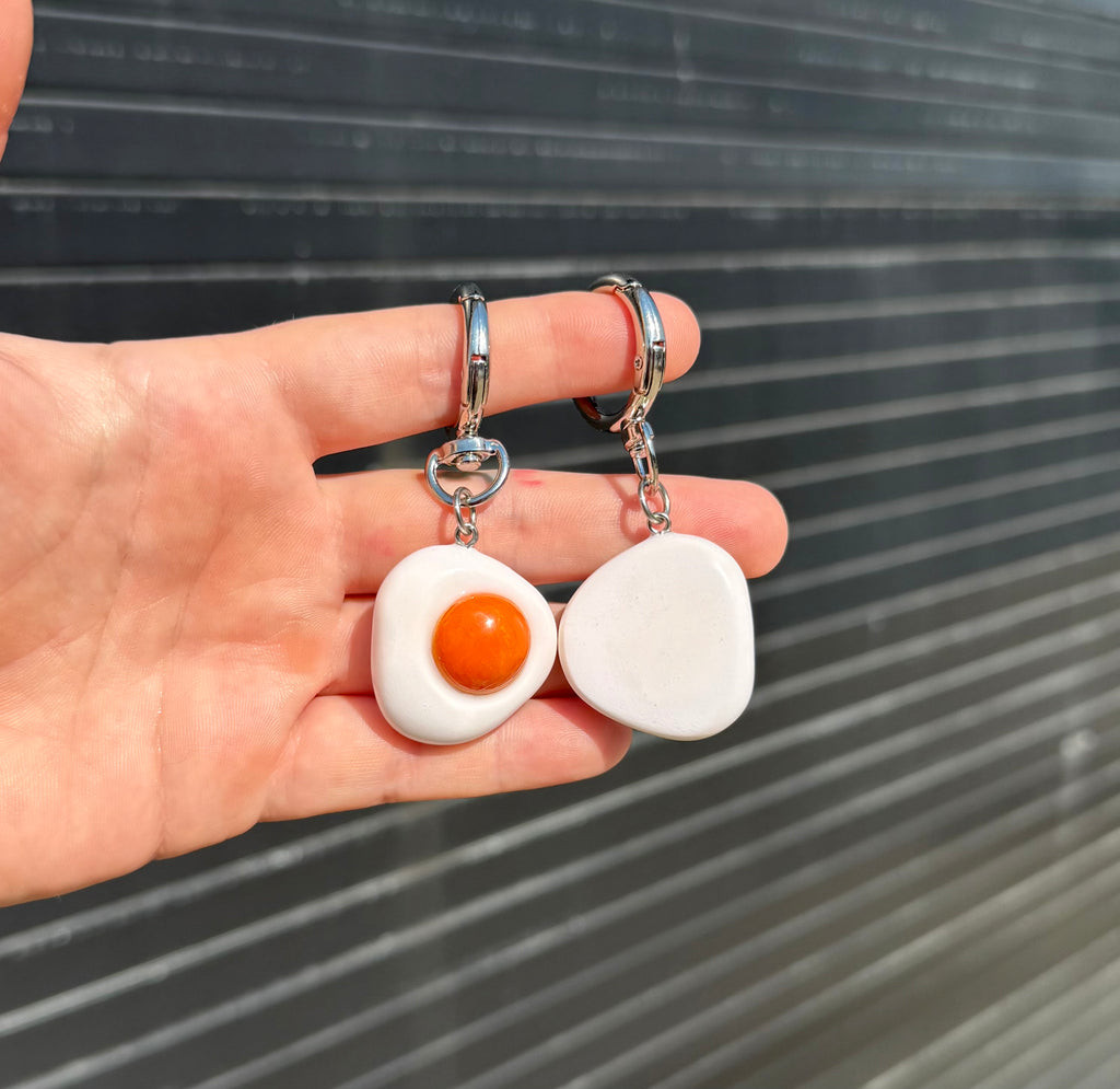 A pair of novelty keychains shaped like fried eggs, held in a hand against a dark corrugated metal background. The earrings feature a white flatbread-textured body with a glossy orange yolk and silver-tone hanger backings.