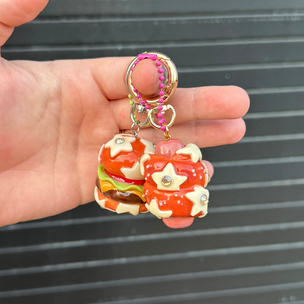 A hand holding a small keychain with two miniature burger charms. The keychain features gold-tone metal rings, a pink braided cord, and two red and white glazed burger charms with small star and heart details, set against a dark gray slatted background.