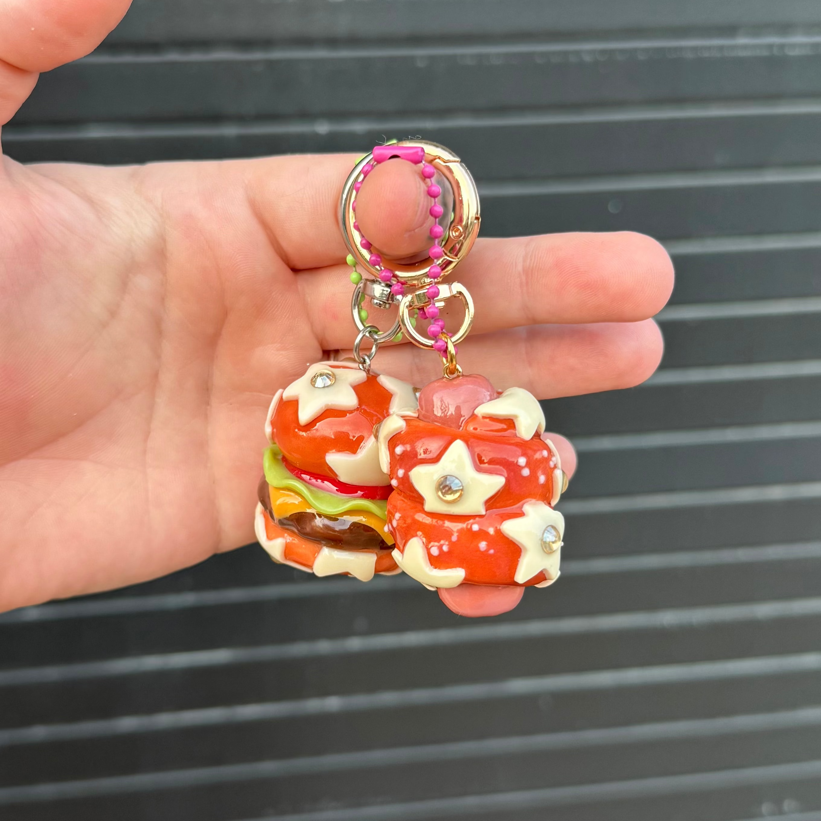 A hand holding a small keychain with two miniature burger charms. The keychain features gold-tone metal rings, a pink braided cord, and two red and white glazed burger charms with small star and heart details, set against a dark gray slatted background.