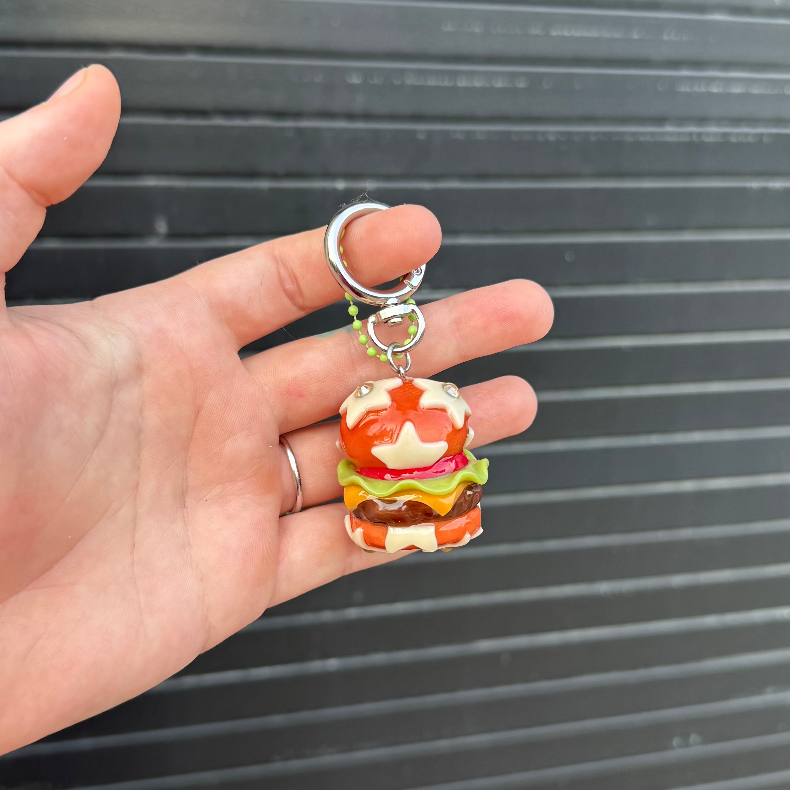 A hand holding a small colorful hamburger keychain with a silver split ring, set against a dark gray corrugated metal background.