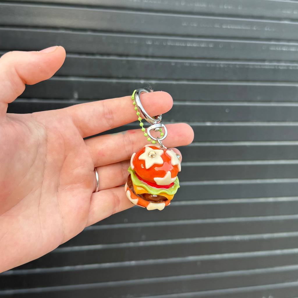 A hand holding a small, colorful polymer clay keychain shaped like a cheeseburger, set against a dark gray corrugated metal background.