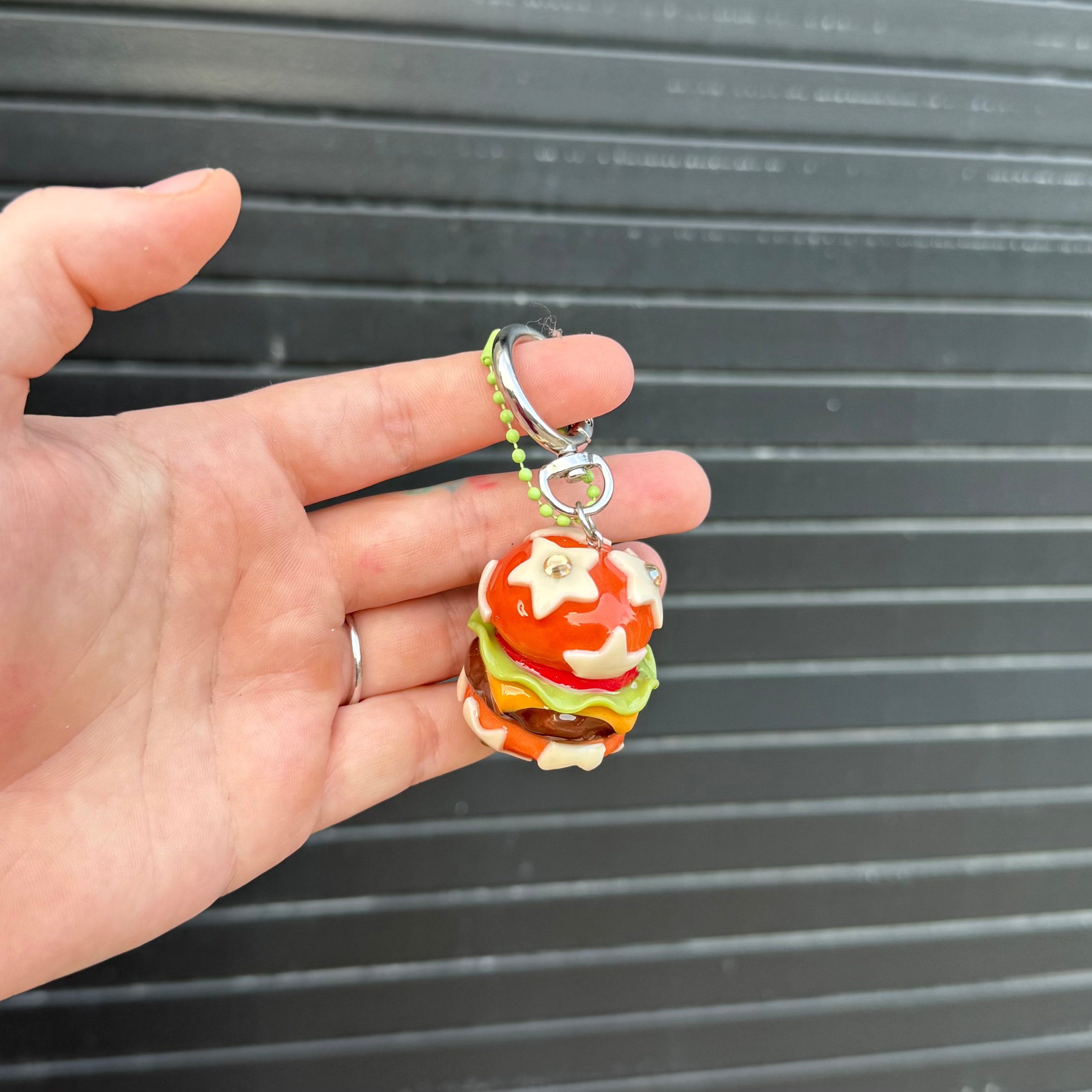 A hand holding a small, colorful polymer clay keychain shaped like a cheeseburger, set against a dark gray corrugated metal background.