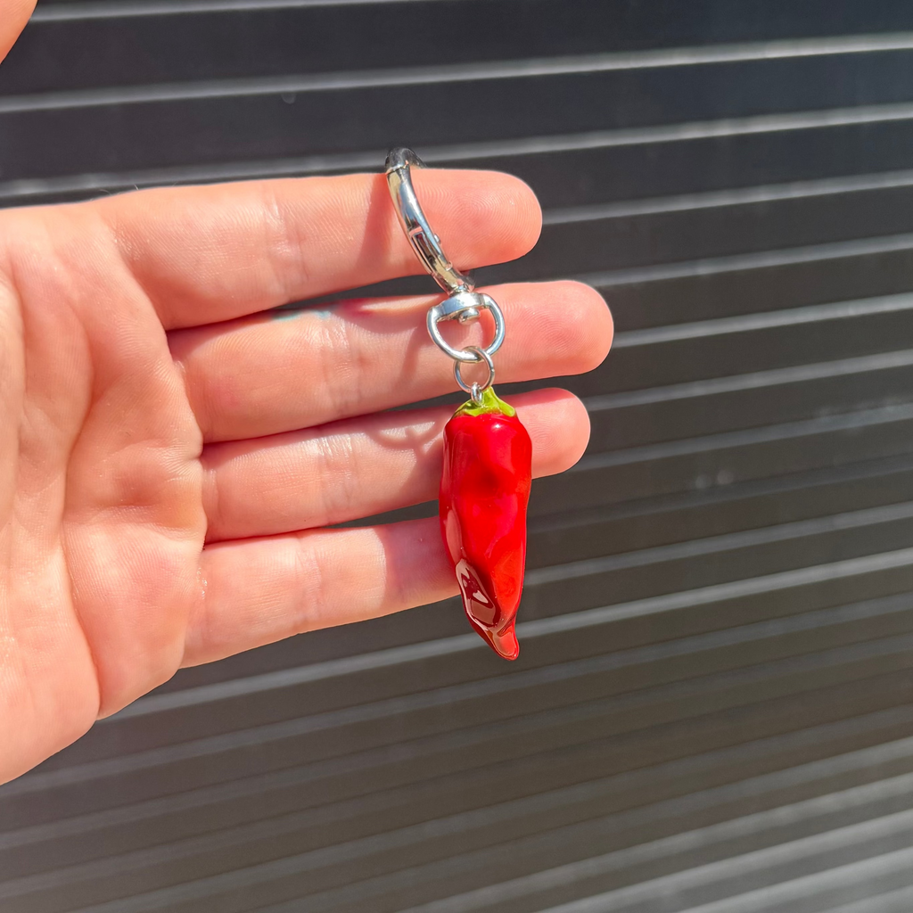 A hand holding a small red chili pepper-shaped keychain with a silver metal clasp against a dark gray corrugated metal background.