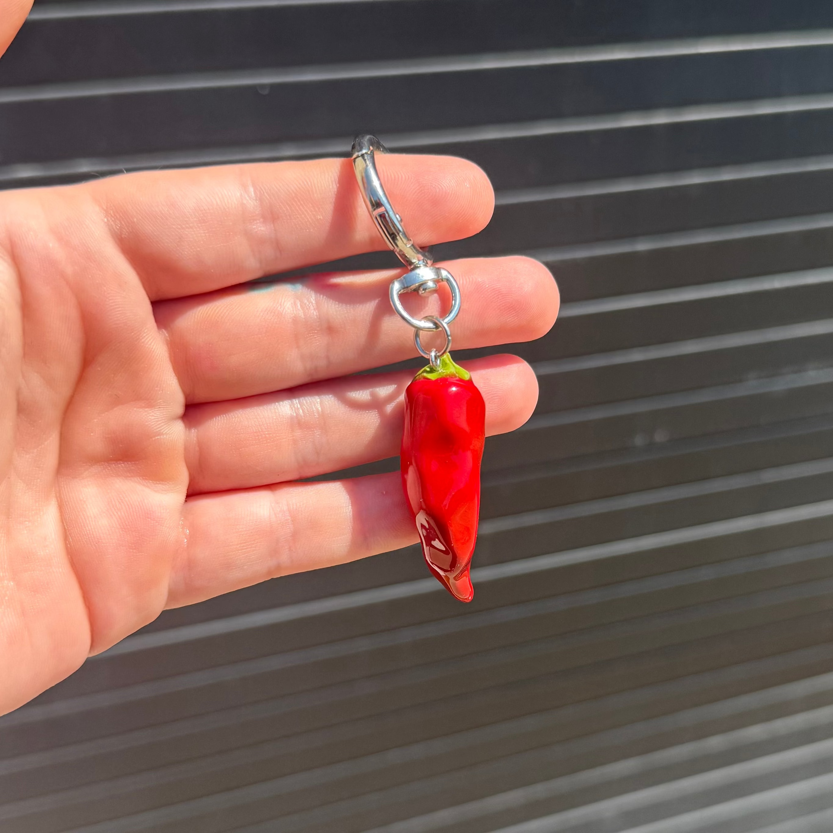 A hand holding a small red chili pepper-shaped keychain with a silver metal clasp against a dark gray corrugated metal background.