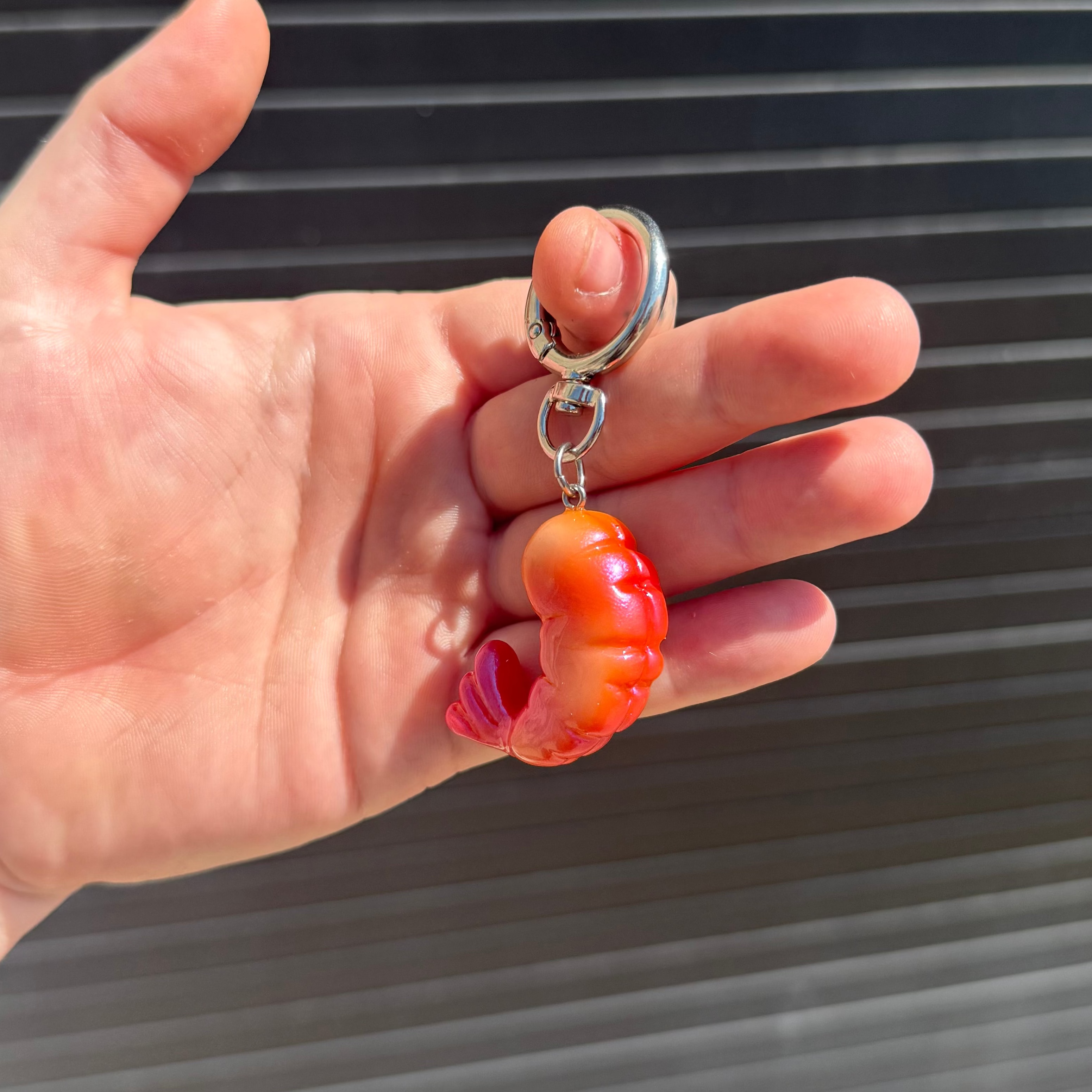 A close-up of a hand holding a small keychain with a realistic orange and red shrimp charm attached to silver metal rings, against a dark corrugated metal background.