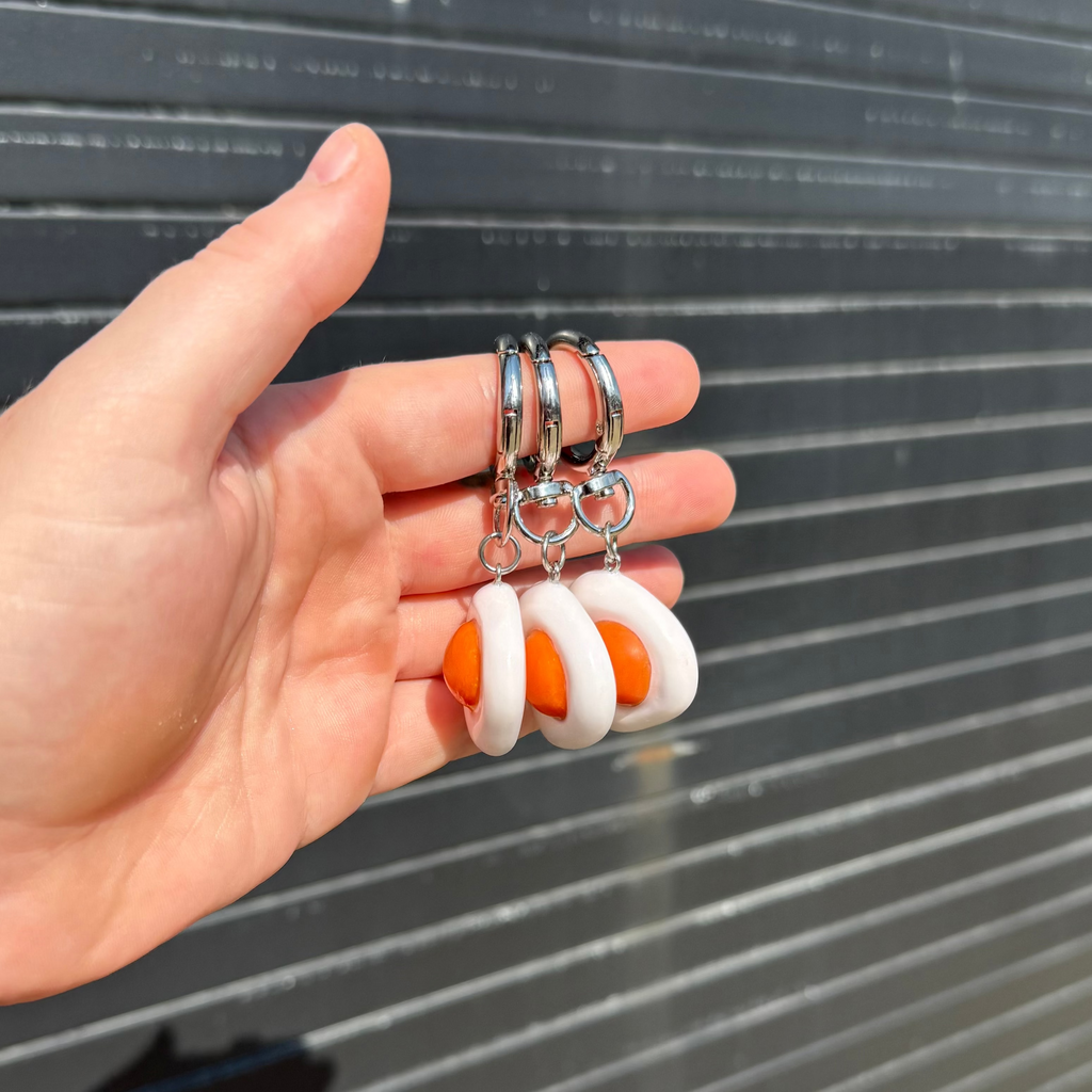 A hand holding a pair of silver hoop earrings with dangling white and orange crescent-shaped charms, set against a dark gray corrugated metal background.