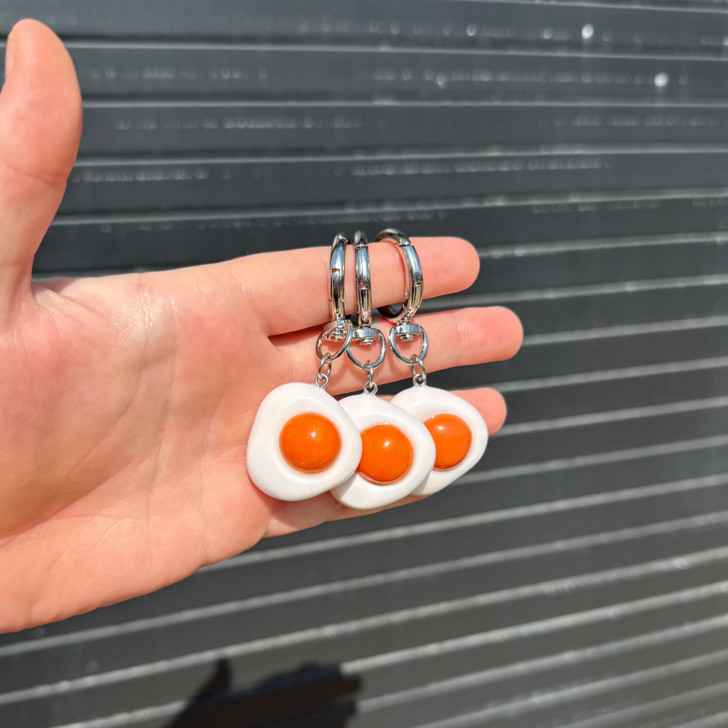 Three mini fried egg keychains with white polymer clay whites and orange yolks on silver metal rings, held in a person's hand against a dark gray slatted background.