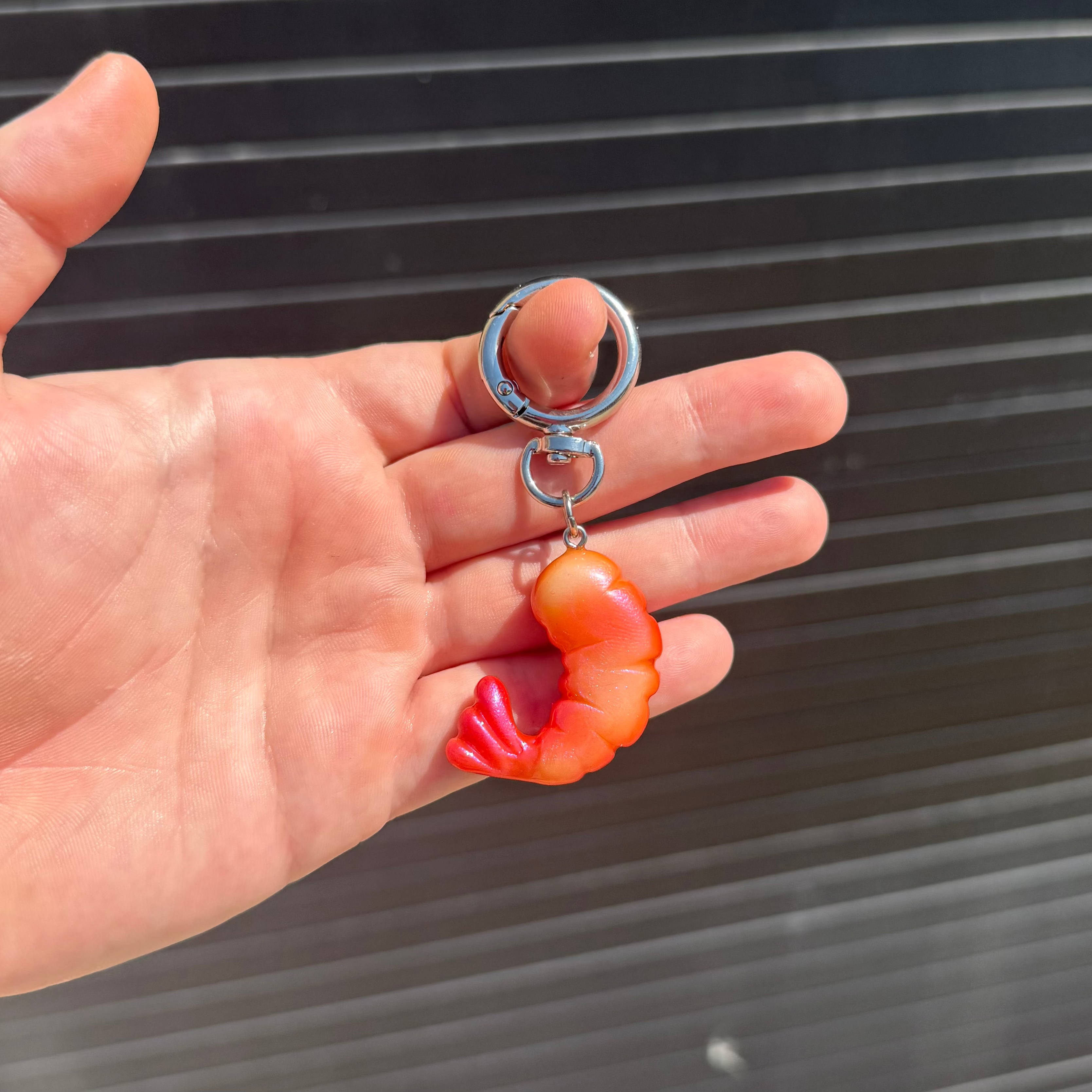 A hand holding a silver split keyring with a dangling shrimp charm in orange and red, set against a dark corrugated metal background.