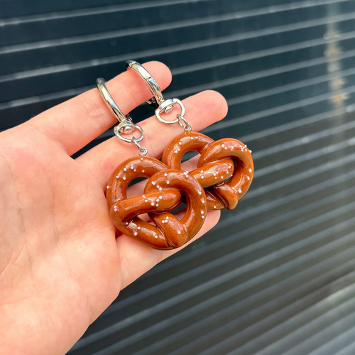 A pair of pretzel-shaped keychains with silver rings, held in a hand against a dark gray slatted background.