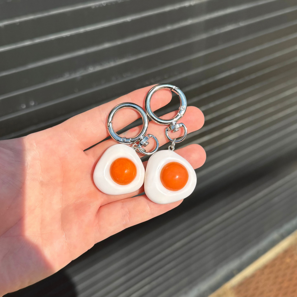 A pair of novelty keychains shaped like fried eggs, held in a person's hand against a dark corrugated metal background. The earrings feature white egg whites, bright orange yolks, and silver-tone metal key rings.