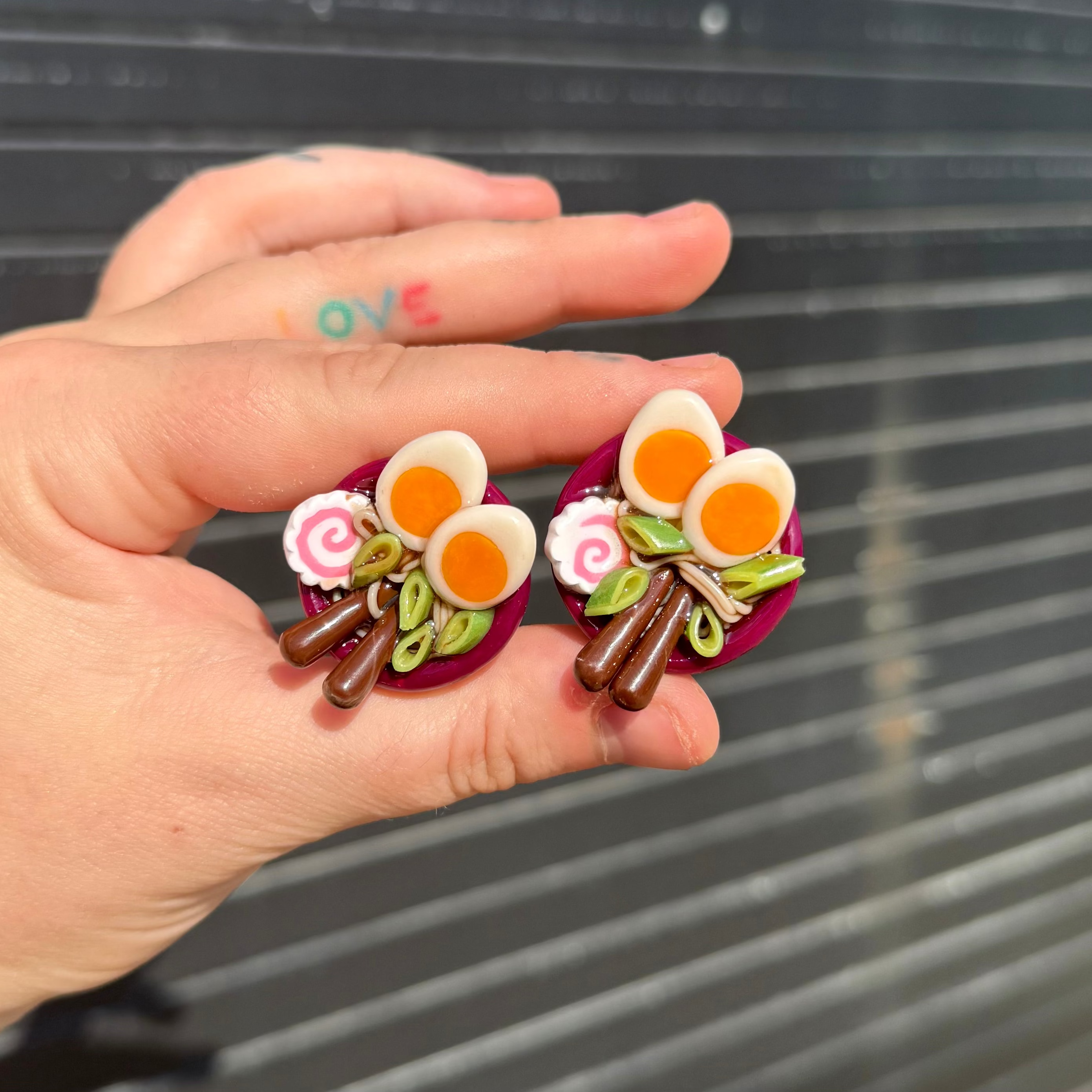 A hand holding a pair of small, colorful, food-themed magnets shaped like ramen bowls with soft-boiled eggs, green onions, and noodles, set against a dark gray slatted background.