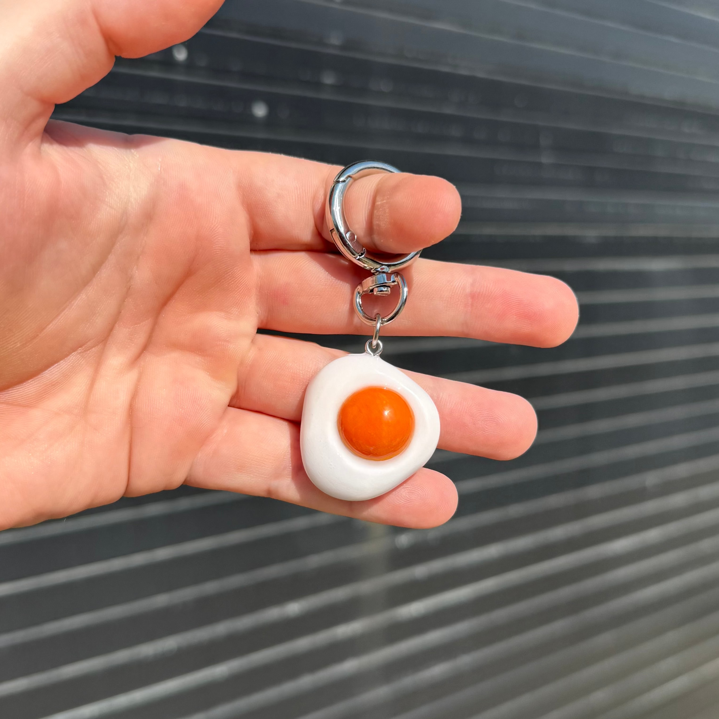 A hand holding a small keychain shaped like a fried egg, with a white oval body, orange yolk, and silver metal rings, against a gray slatted background.