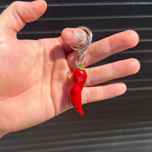 A hand holding a small red chili pepper keychain with a silver metal split ring and keychain clips against a dark gray slatted background.