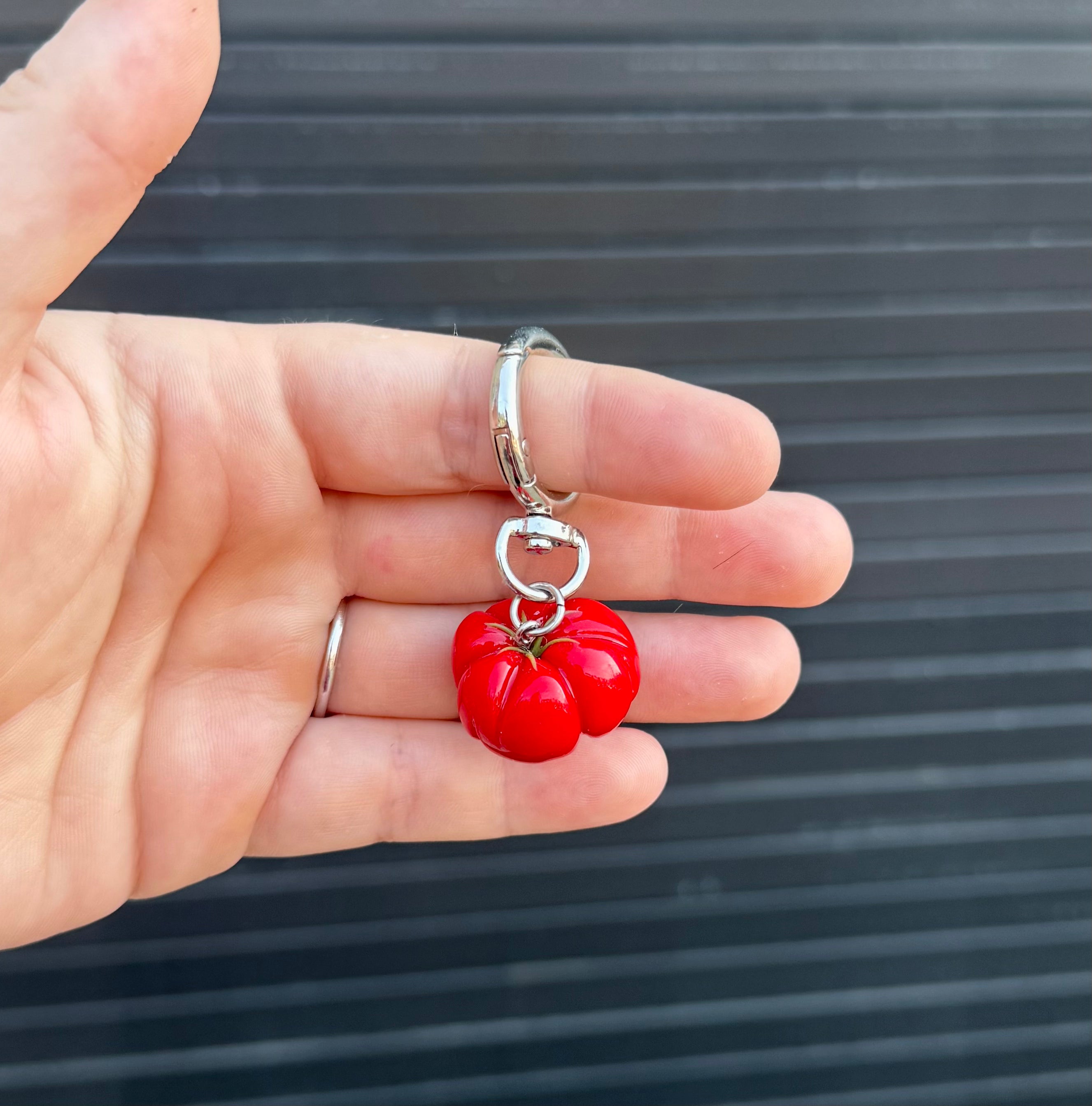 A hand holding a silver split ring keychain with a glossy red tomato charm against a dark gray slatted background.