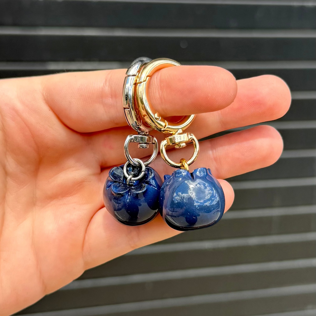 a hand holding two blueberry dangle keychains in front of a graphite back drop. 