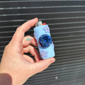 A hand holding a light blue polymer clay pocket lighter decorated with purple star stickers and a dark blue blueberry, set against a dark gray slatted background.
