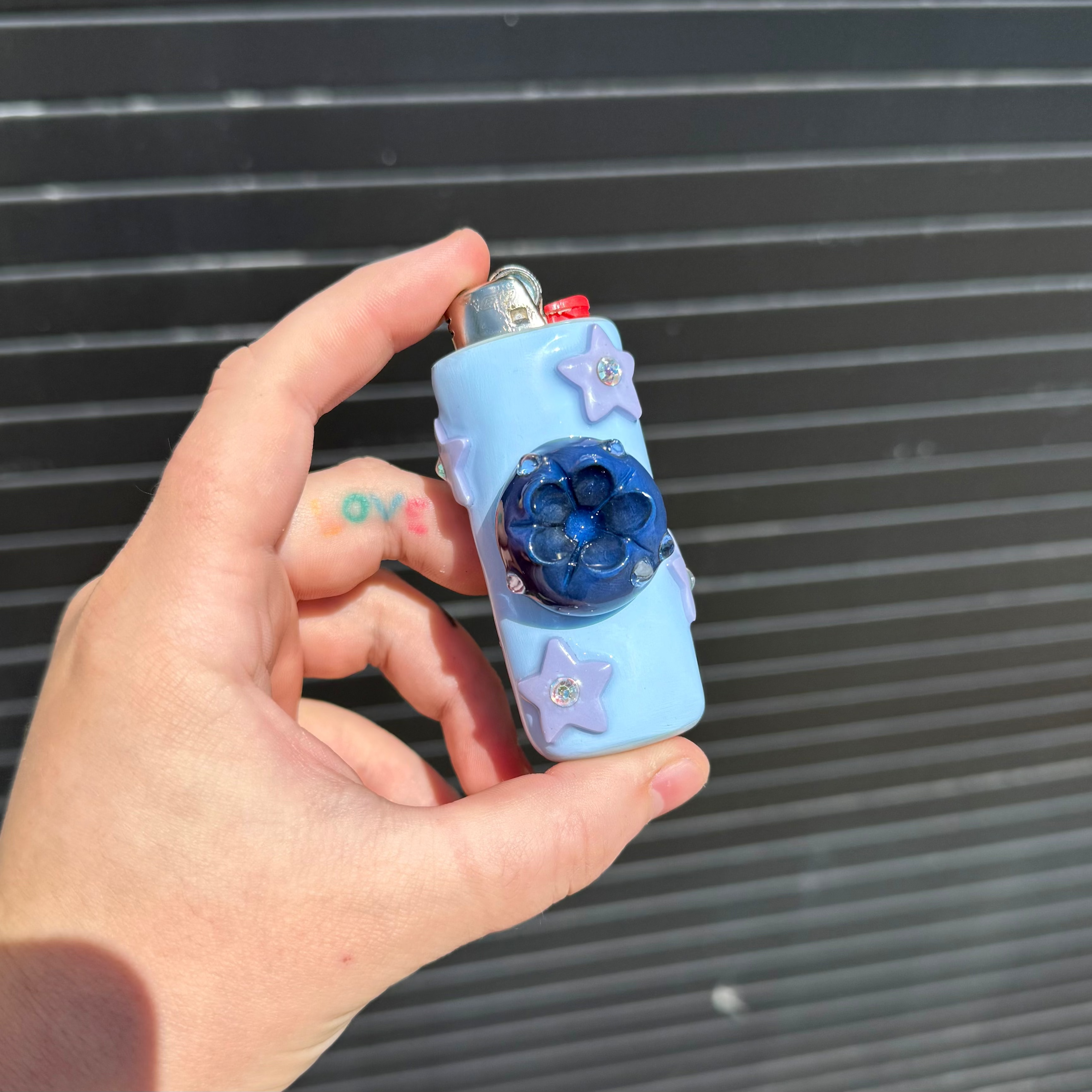A hand holding a light blue polymer clay pocket lighter decorated with purple star stickers and a dark blue blueberry, set against a dark gray slatted background.
