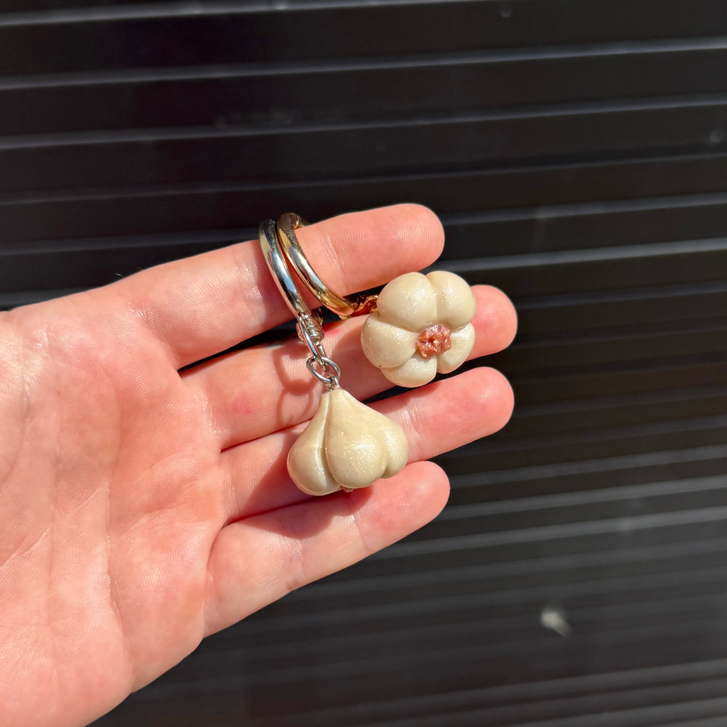A hand holding a pair of garlic-shaped keychains with silver and rose gold rings, set against a dark corrugated metal background.