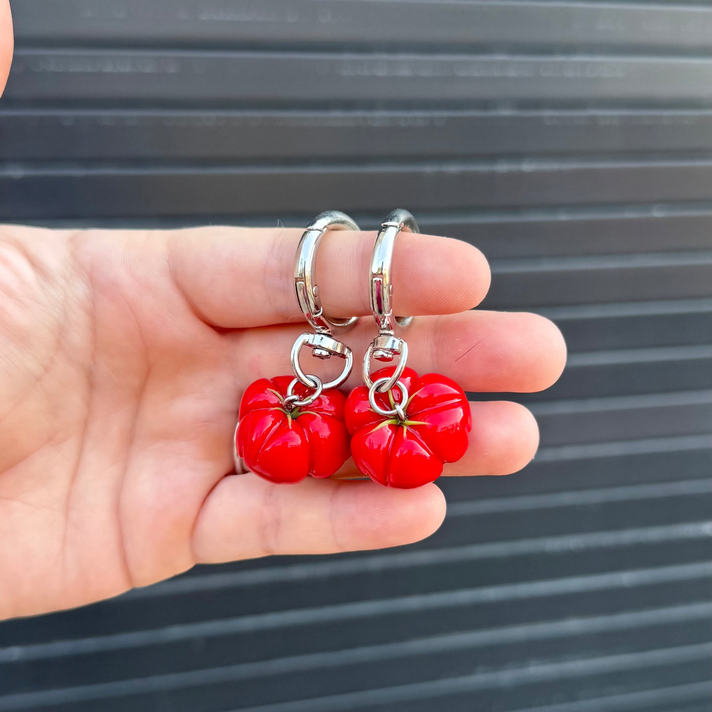 A pair of red heirloom tomato keychains with silver metal rings, held in a person's hand against a dark gray corrugated metal background.