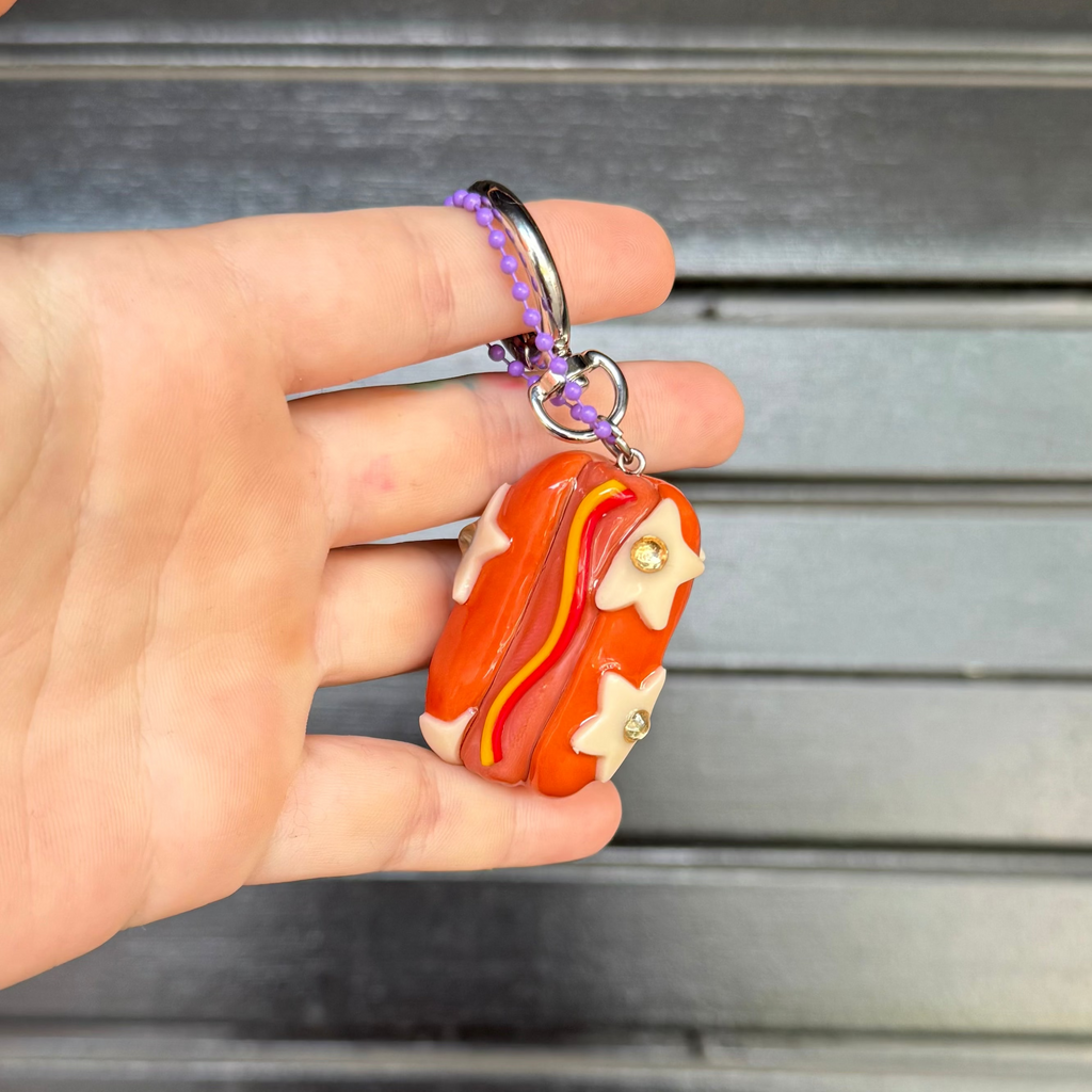 A hand holding a small orange and red hotdog keychain charm with cream star details, set against a dark gray slatted background.