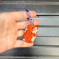 A hand holding a small orange and red hotdog keychain charm with cream star details, set against a dark gray slatted background.