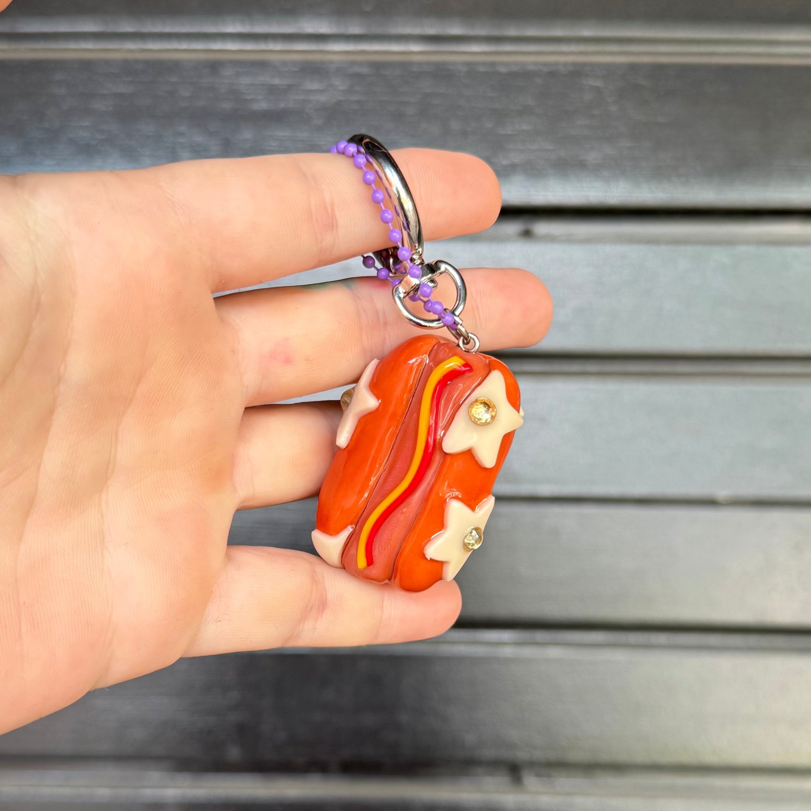 A hand holding a small orange and red hotdog keychain charm with cream star details, set against a dark gray slatted background.