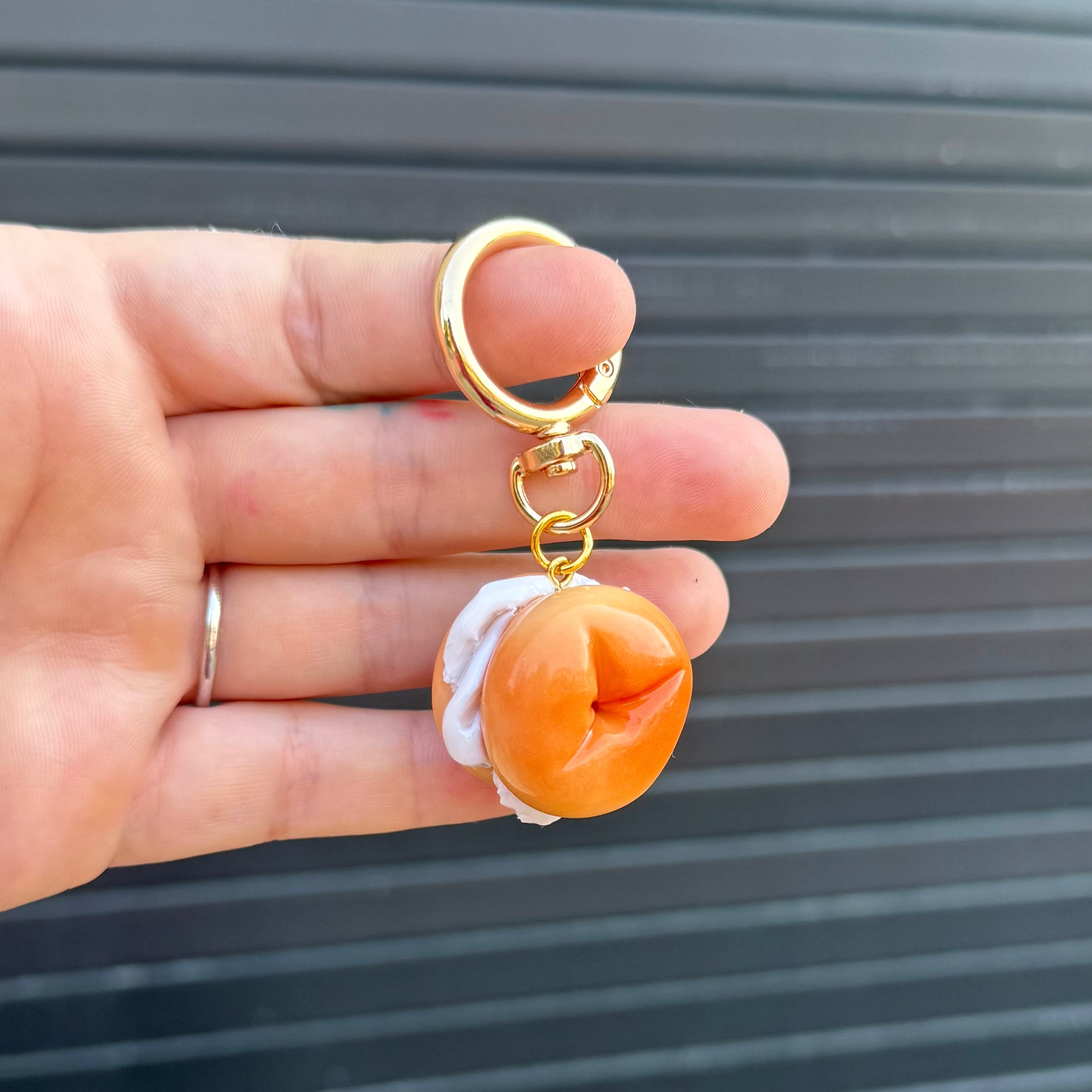 A hand holding a novelty keychain featuring a small bagel charm, against a dark gray slatted background.