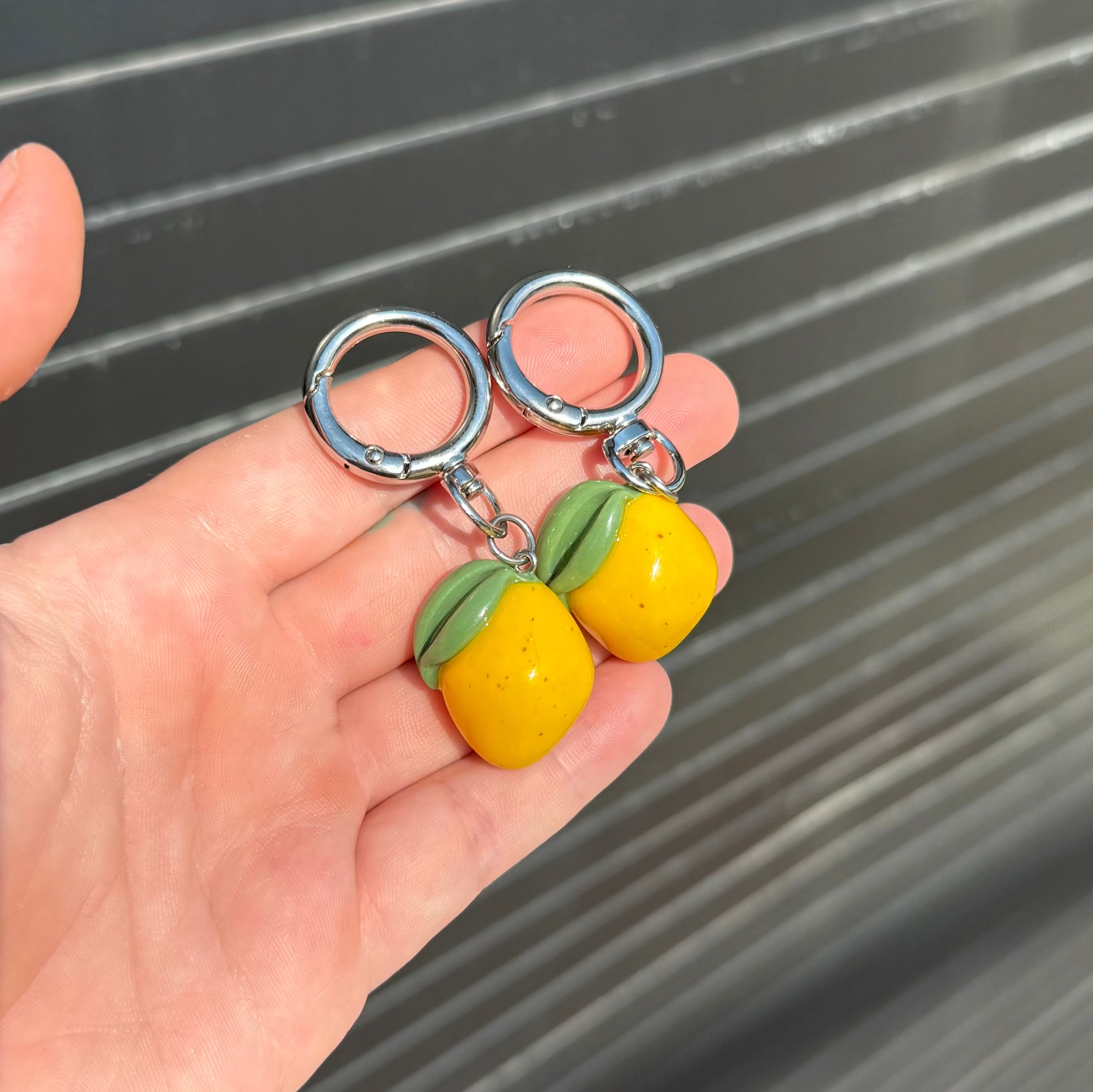 Two small yellow fruit charms with green tops attached to silver key rings, held in a person's hand against a gray slatted background.