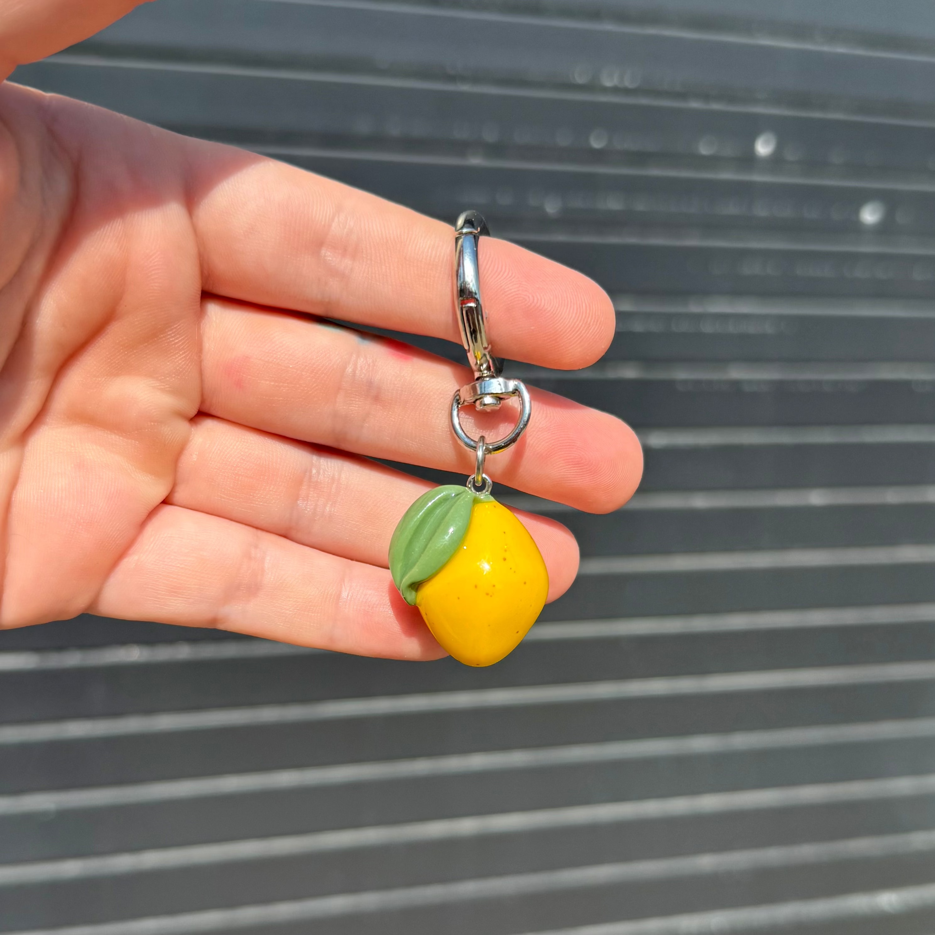 A hand holding a small yellow lemon-shaped charm with a green leaf detail and a silver metal clasp, set against a dark gray slatted background.