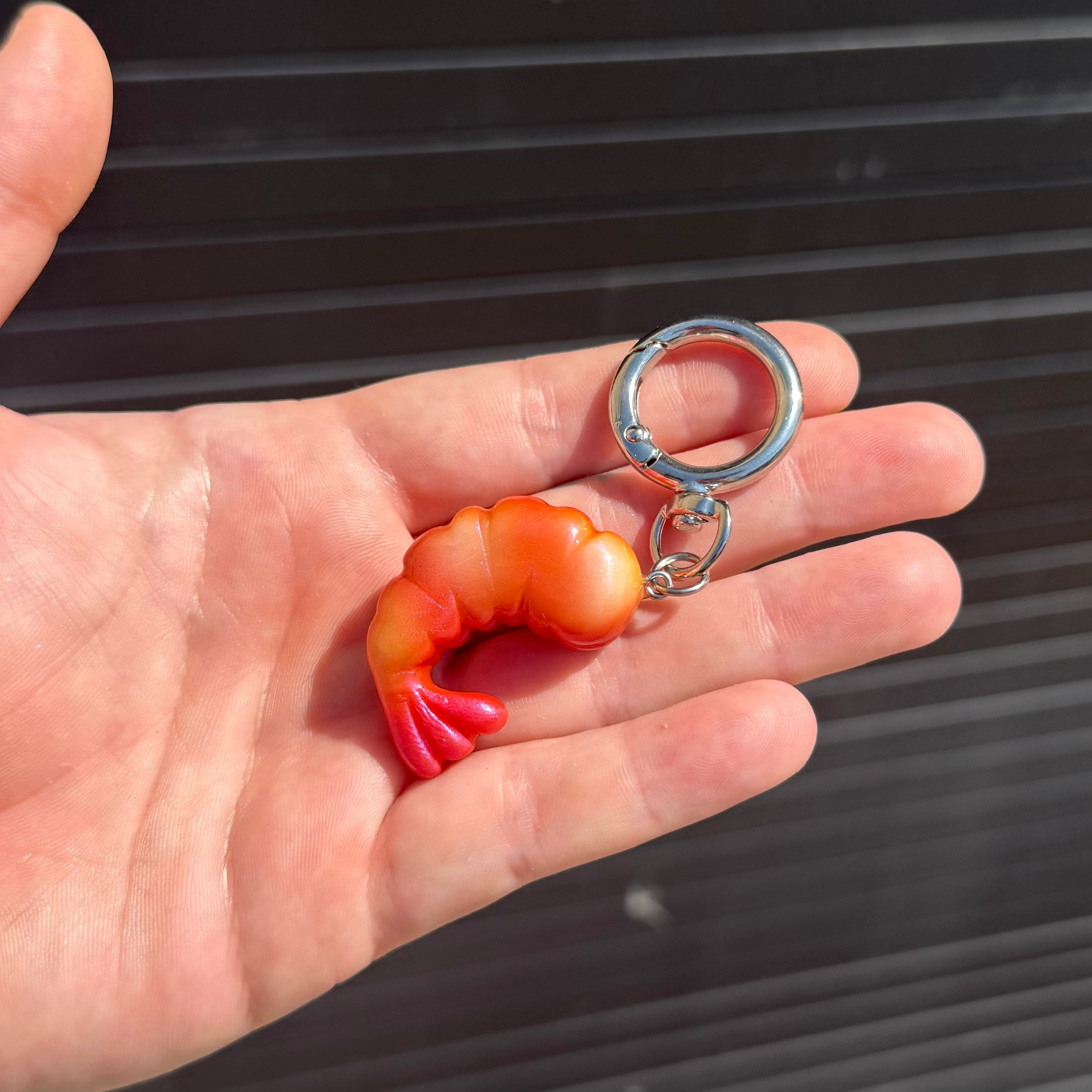 A small shrimp-shaped keychain in orange and red held in a person's hand, with a silver metal key ring and split ring against a dark slatted background.