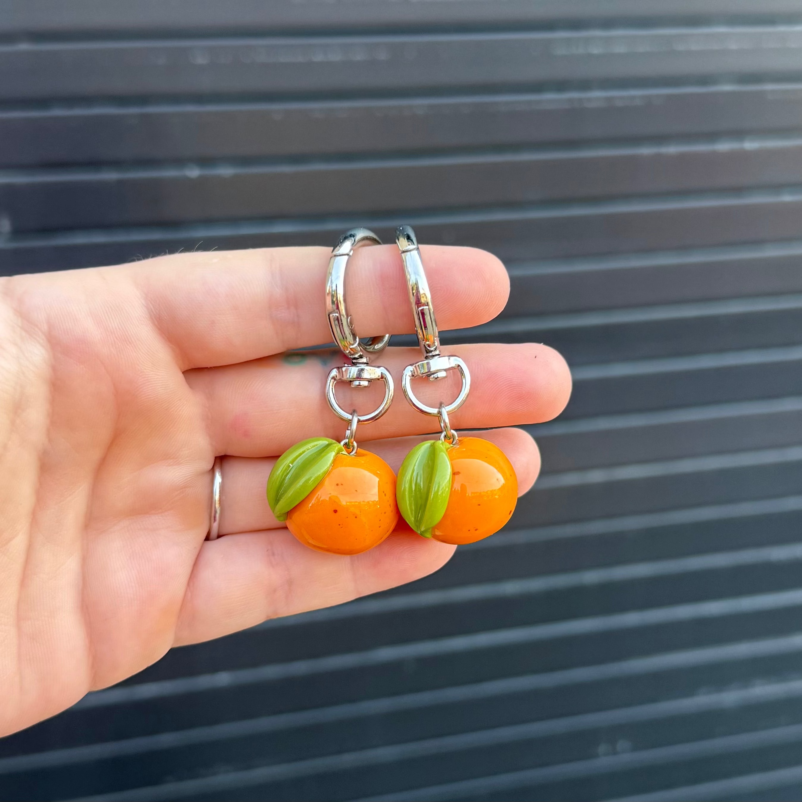 A pair of keychains with dangling orange citrus-shaped charms and green leaf accents, held in a person's hand against a dark gray slatted background.