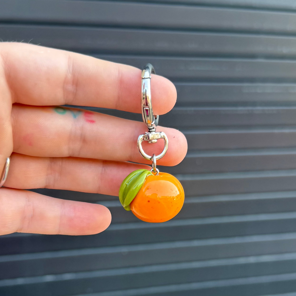 orange fruit keychain charm being held against a graphite colored wall. 