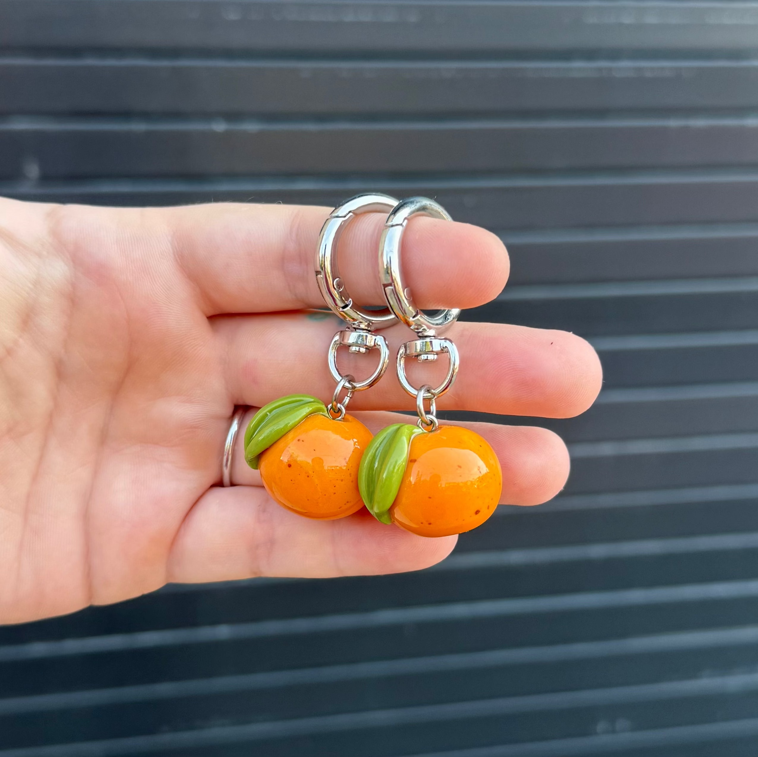 A pair of novelty keychains shaped like glossy orange fruits with green leaves, held in a person's hand against a dark gray slatted background.