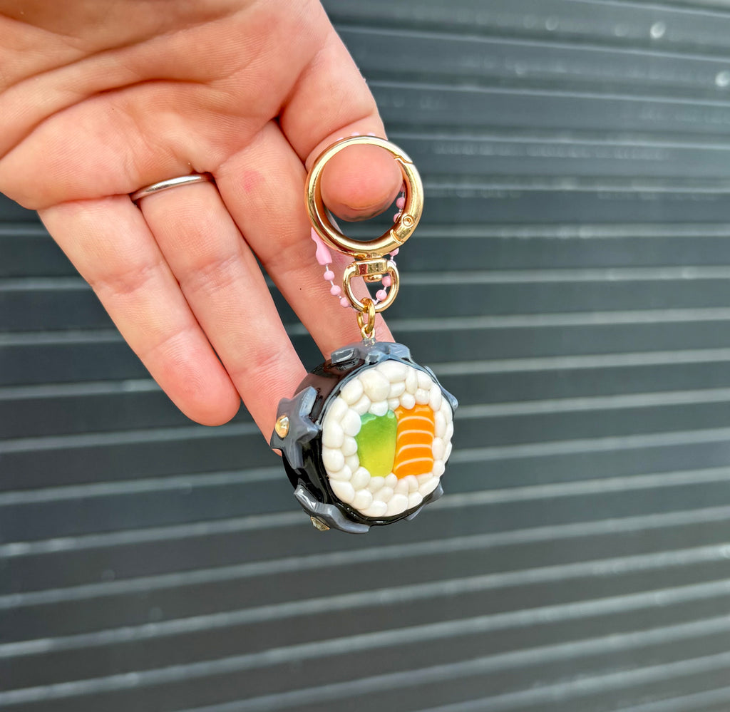 A hand holding a sushi-themed keychain against a dark gray slatted background. The keychain features a detailed maki sushi charm with white rice, green and orange filling, and a gold key ring.