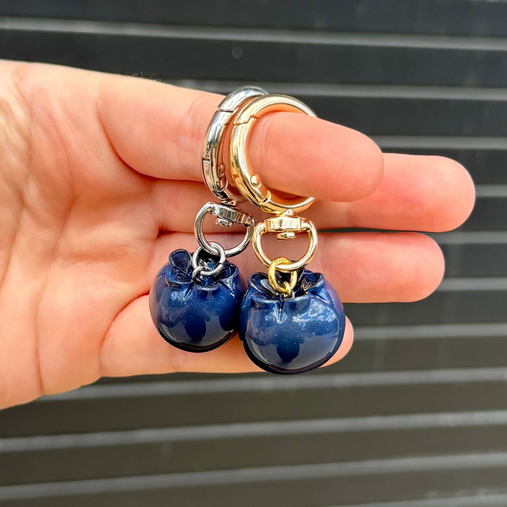 A hand holding two small blueberry keychains with gold and silver metal rings, set against a dark gray slatted background.