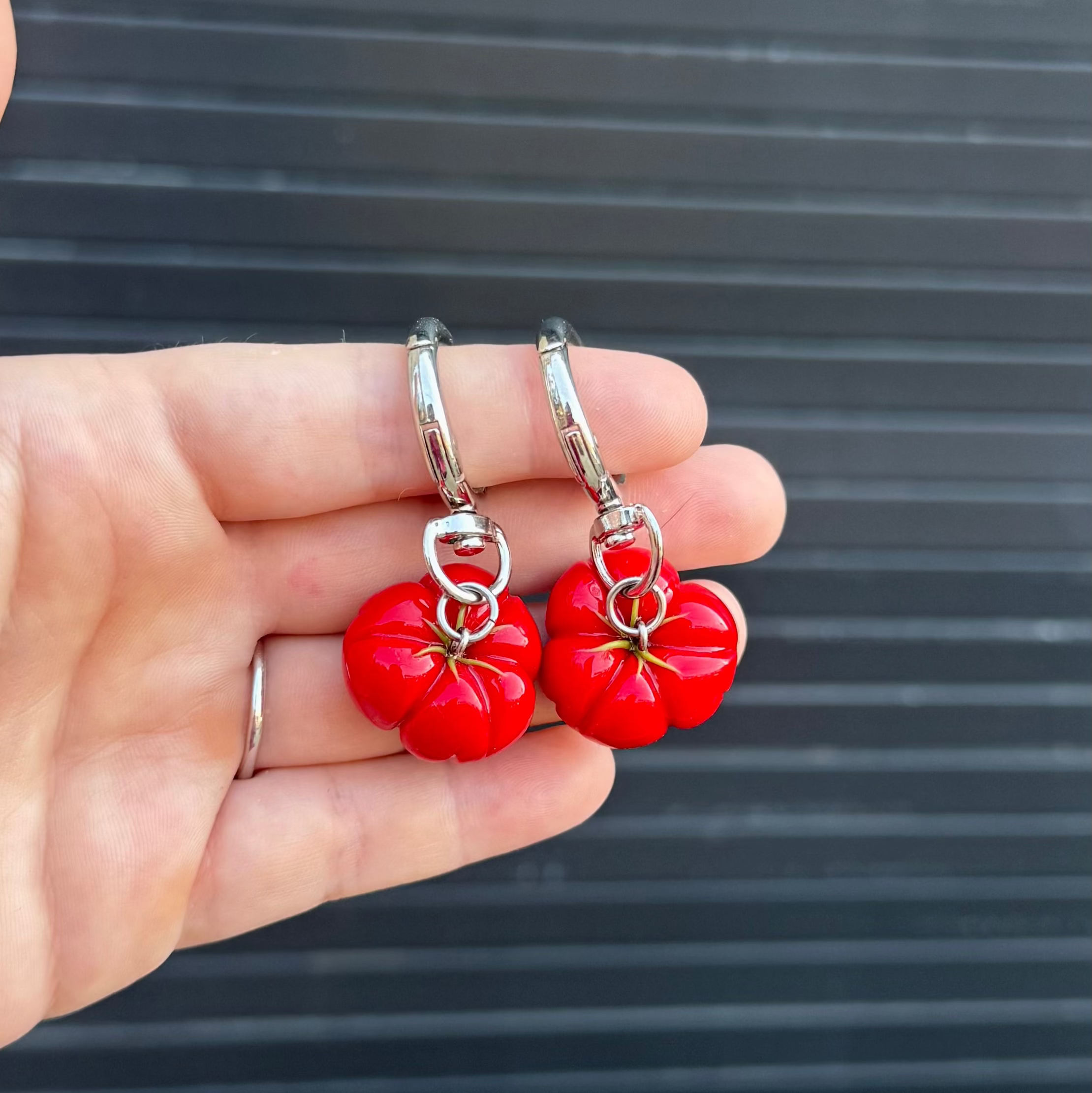 A pair of silver keychains with dangling red tomato charms, held in a person's hand against a dark gray slatted background.