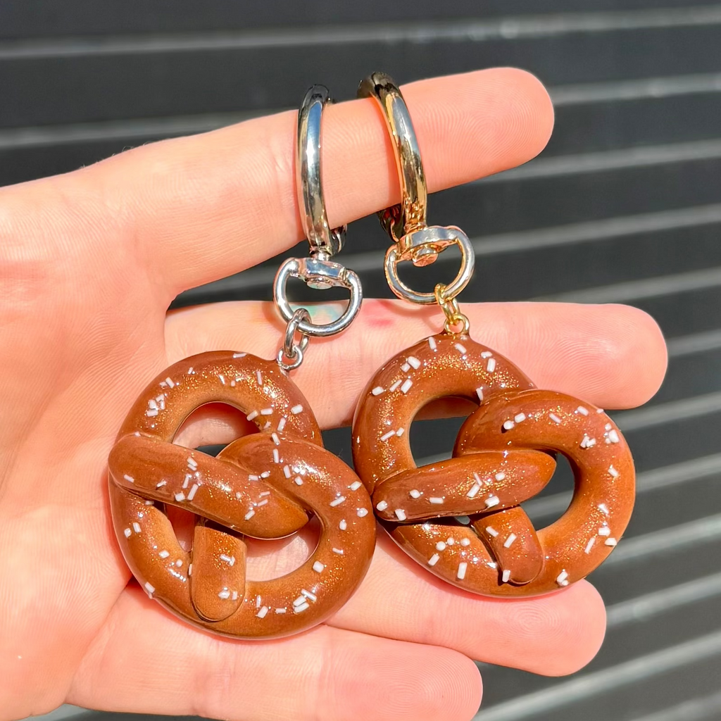 A pair of novelty keychains shaped like pretzels, held in a hand. The pretzel charms are brown with white salt details, attached to gold and silver fishhook ear wires.