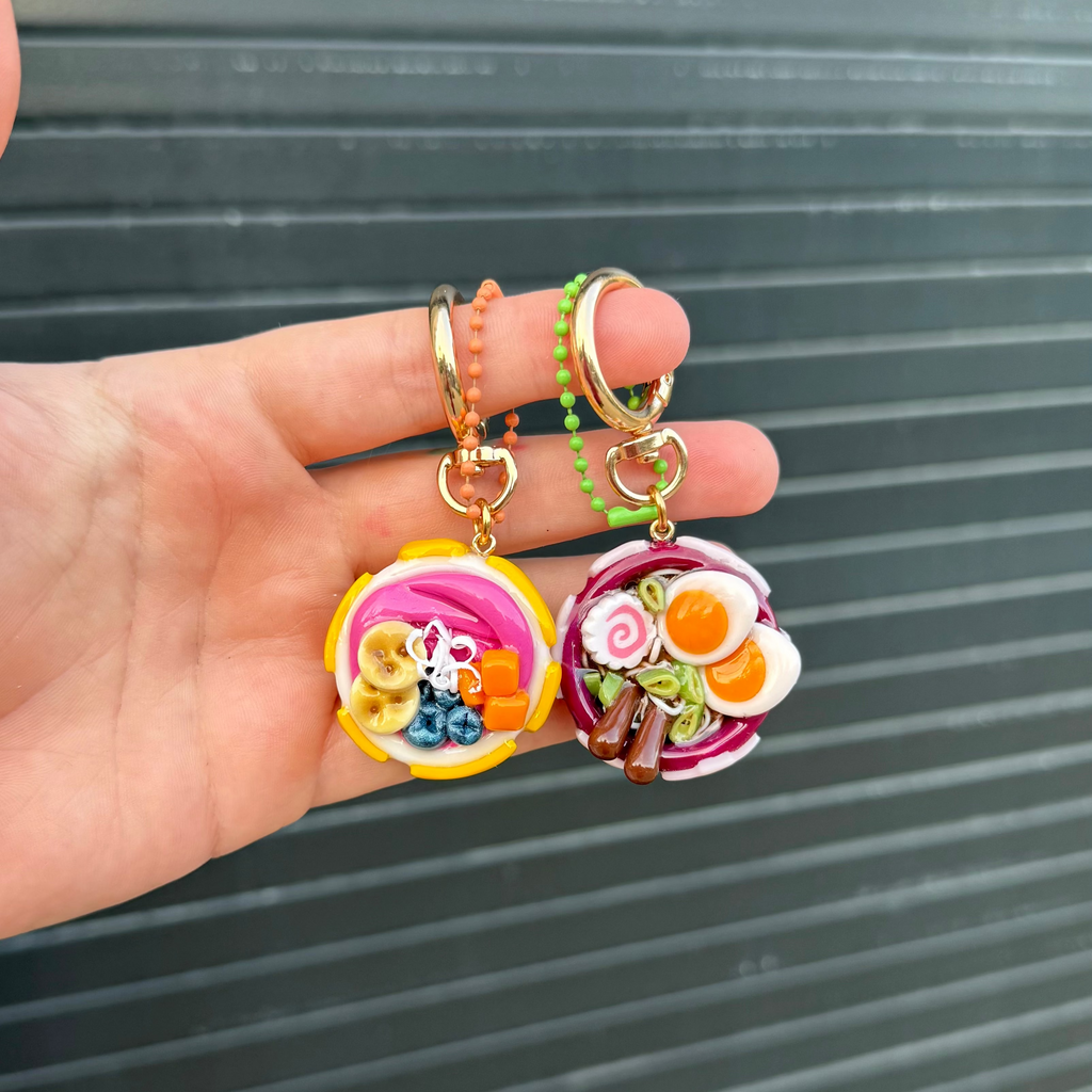 A hand holds two small food-themed keychains against a dark gray slatted background. One keychain features a white bowl with yellow stars holding a acai smoothie with banana slices, mango chunks, coconut shreds and blueberries. The other features a purple ramen bowl design with a fish chip, chopsticks, green onion and eggs.