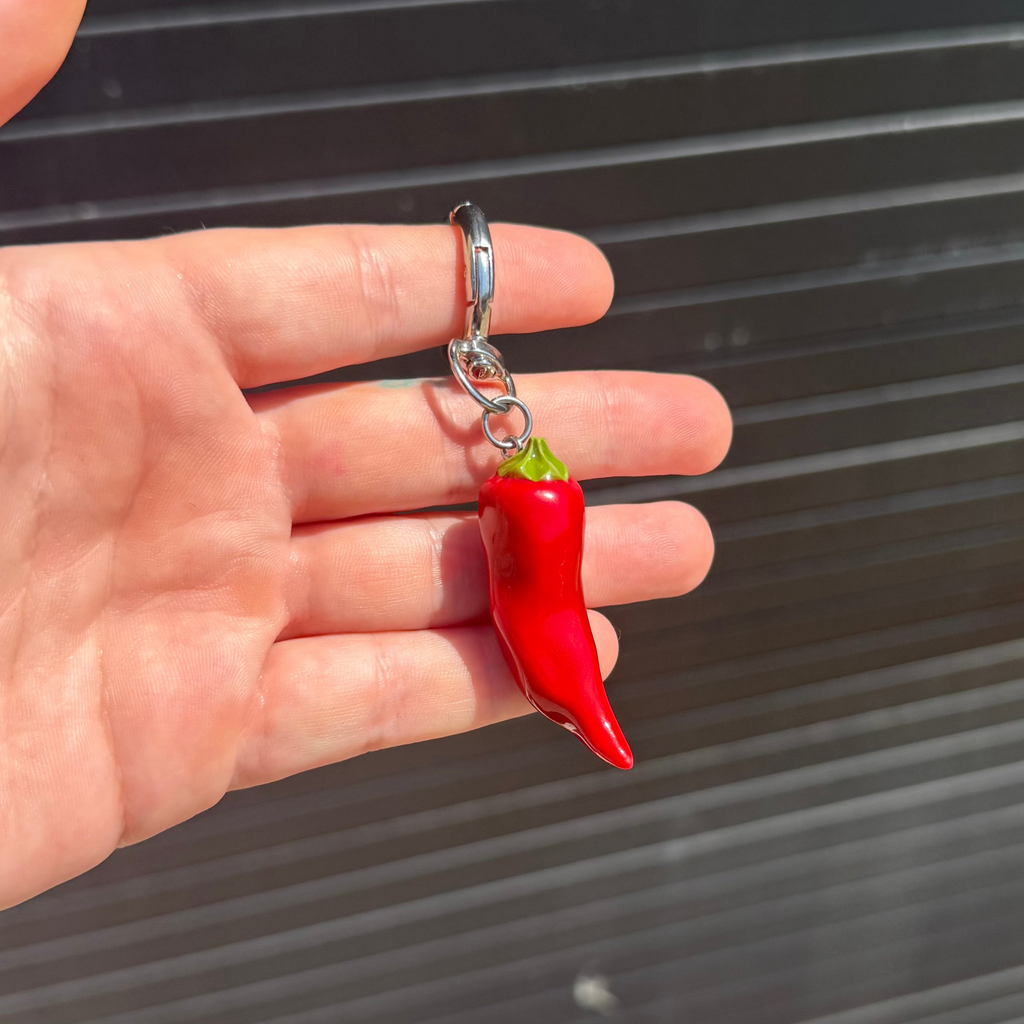 A hand holding a small red chili pepper keychain with a silver metal ring and chain against a dark gray slatted background.