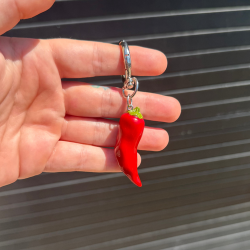 A hand holding a small red chili pepper keychain charm with a silver metal split ring and clasp, set against a dark gray corrugated metal background.