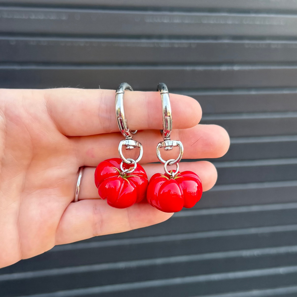 A hand holding a pair of silver keychains with red glossy tomato-shaped charms, set against a dark gray corrugated metal background.