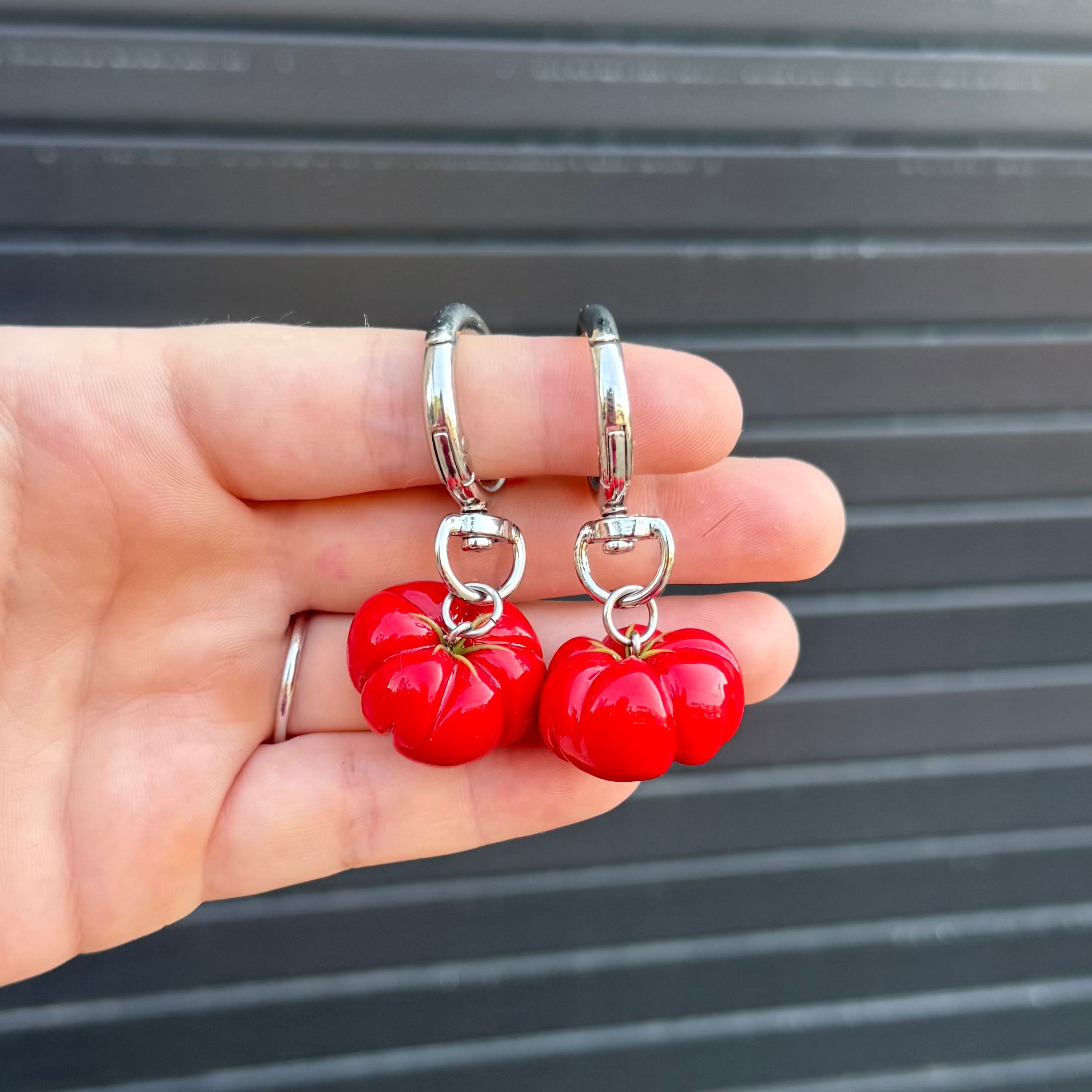 A hand holding a pair of silver keychains with red glossy tomato-shaped charms, set against a dark gray corrugated metal background.