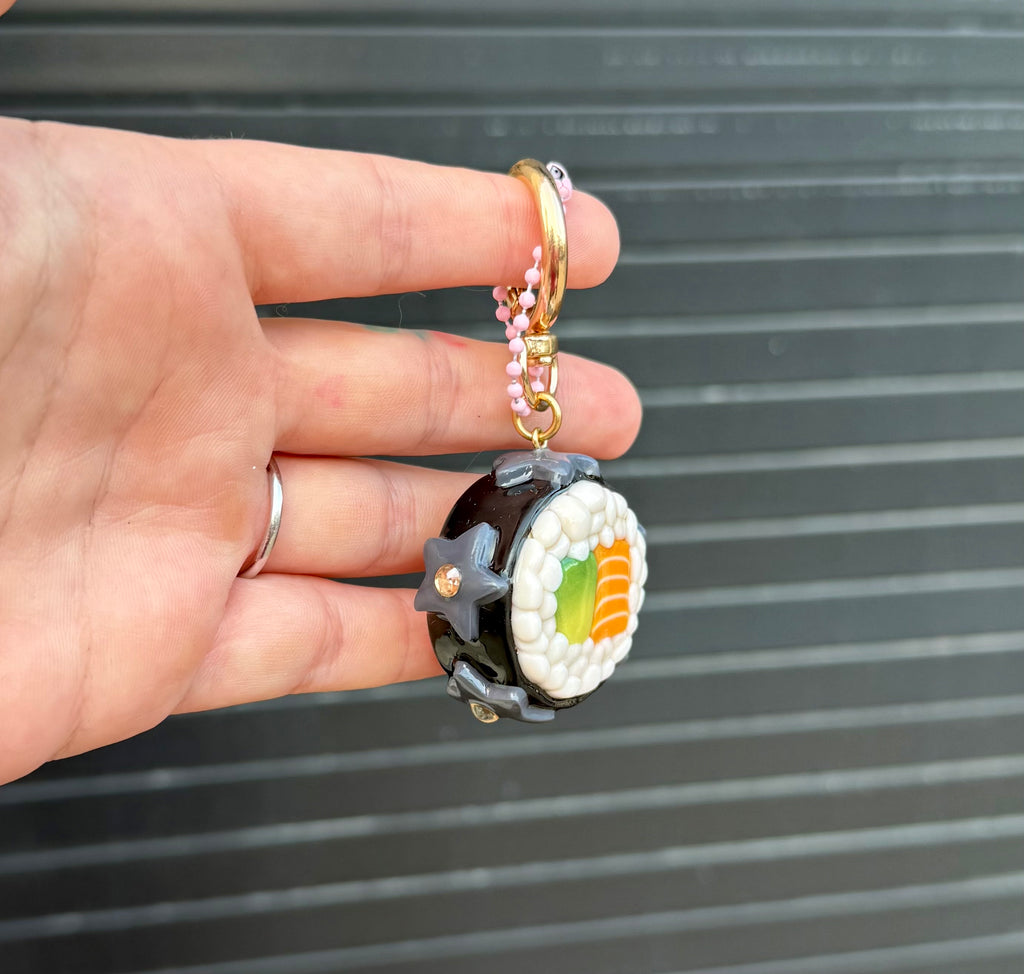 A hand holding a small sushi-themed keychain or charm against a dark gray slatted background. The charm is shaped like a maki roll, with white rice, green and orange filling details, a dark outer ring, and a gold key ring and chain.