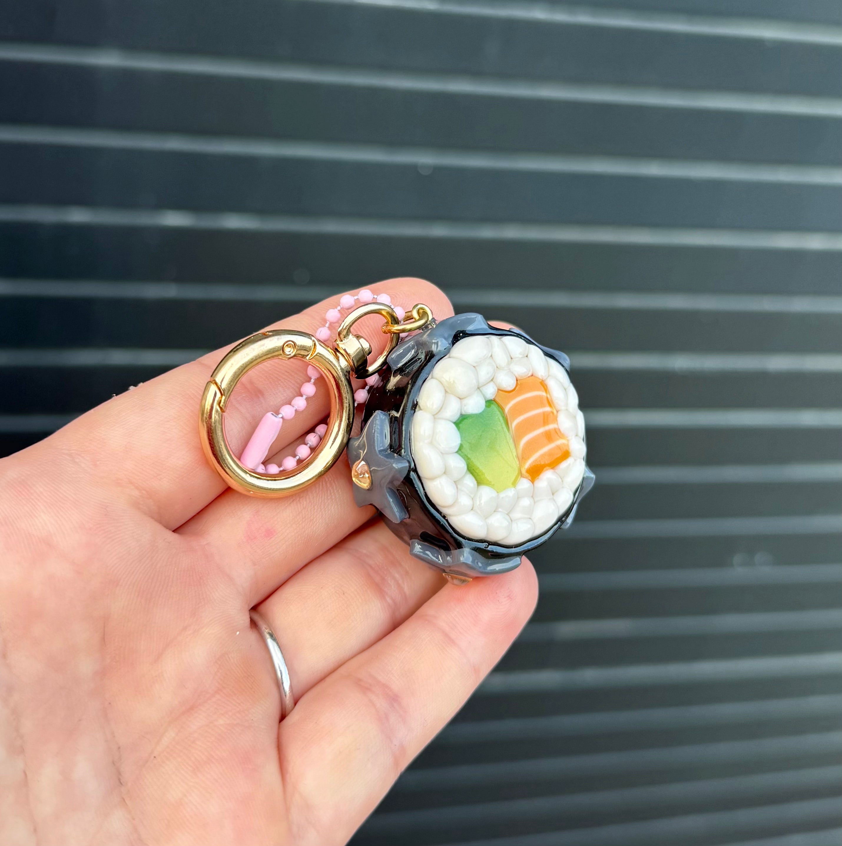 A hand holding a small sushi-themed keychain charm with a gold ring and pink beaded strap, against a dark gray slatted background.